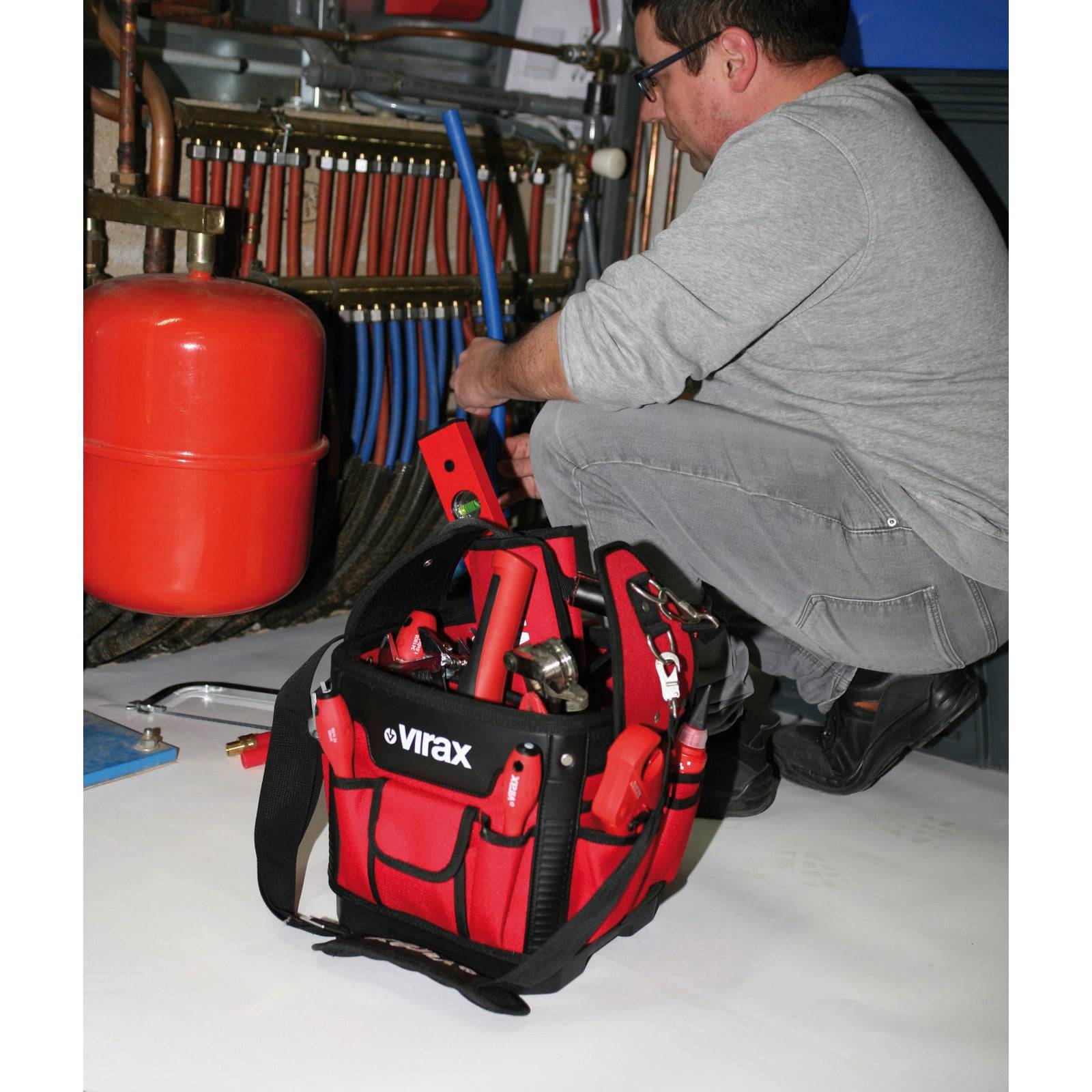Eine Person kniet vor Rohrleitungen und benutzt ein Werkzeug aus einer roten Werkzeugkiste. Ein roter Drucktank ist im Hintergrund sichtbar.