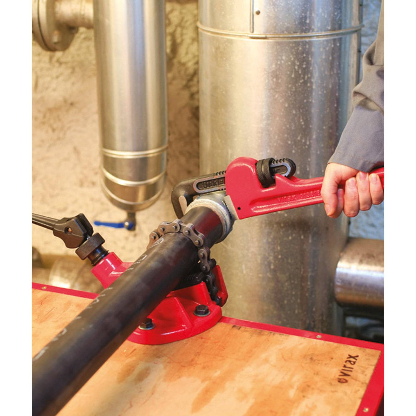 Eine Person, die einen roten Rohrzange auf einem großen schwarzen Rohr in einer industriellen Umgebung verwendet, umgeben von Metallrohren und einer Holzoberfläche.