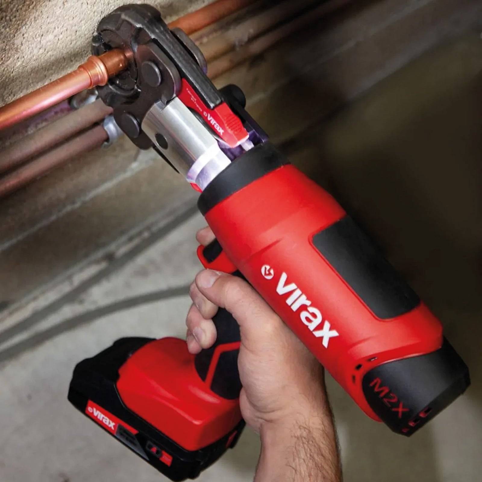 Eine Hand verwendet ein rotes und schwarzes Virax M2X-Werkzeug, um ein Kupferrohr an einer Wand zu befestigen, was die Anwendung des Werkzeugs bei Installationsarbeiten verdeutlicht.