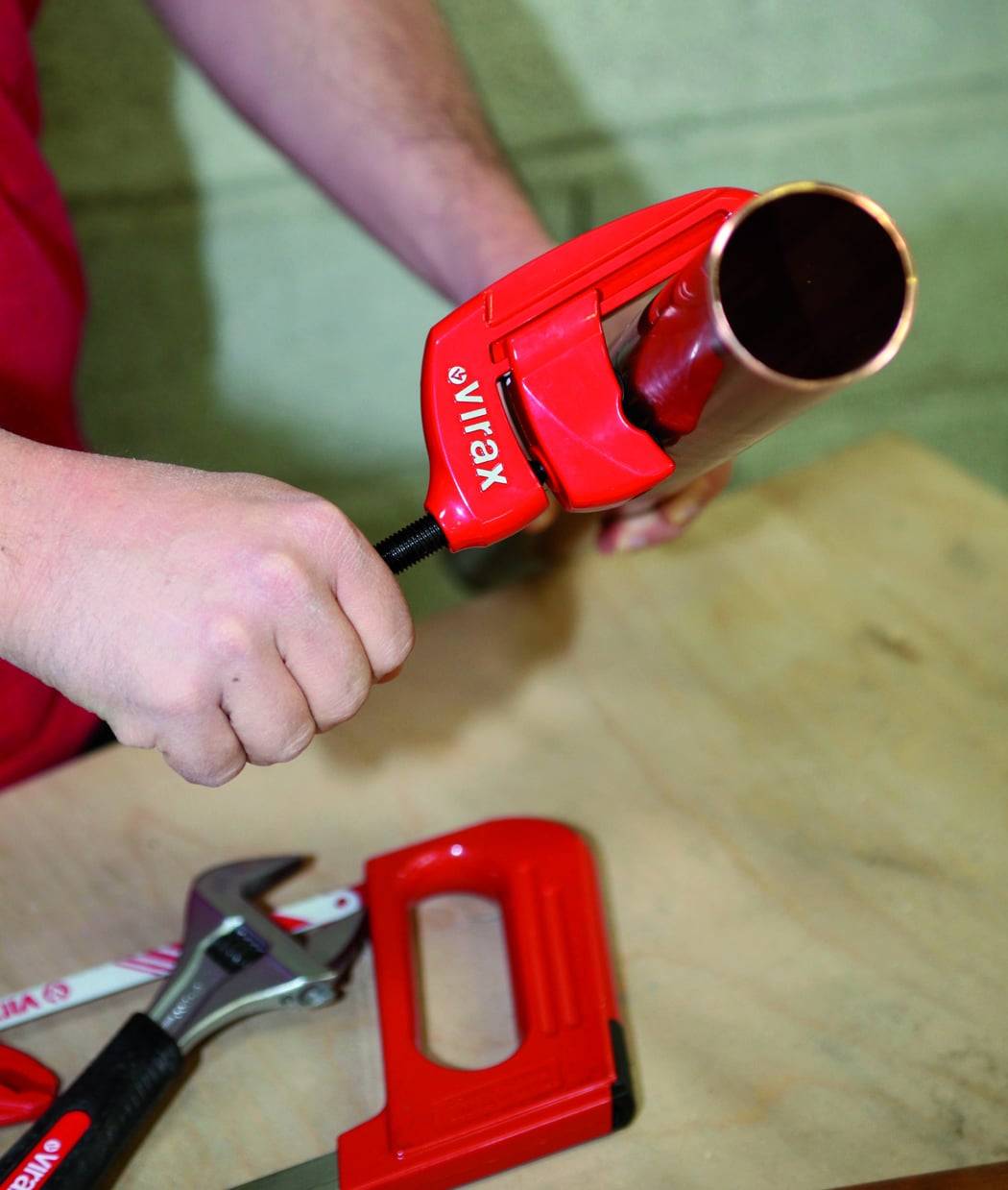 Eine Person in einem roten Hemd benutzt einen roten Rohrabschneider auf einem Kupferrohr, wobei ein Schraubenschlüssel und ein Teil einer Säge auf dem Tisch sichtbar sind.