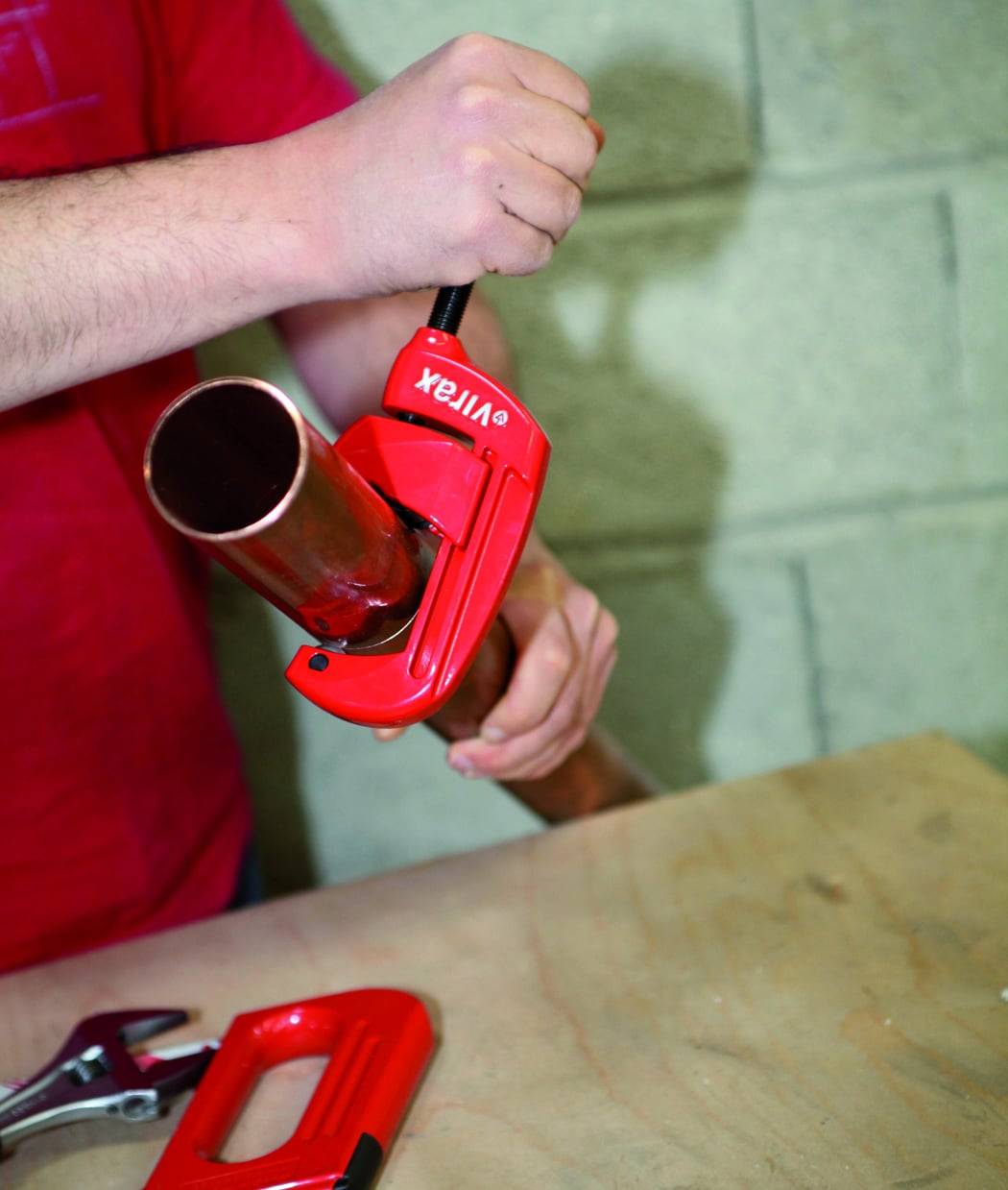 Eine Hand, die einen roten Rohrabschneider auf einem Kupferrohr verwendet, vor einem unscharfen Hintergrund aus Betonwand und Holzoberfläche.