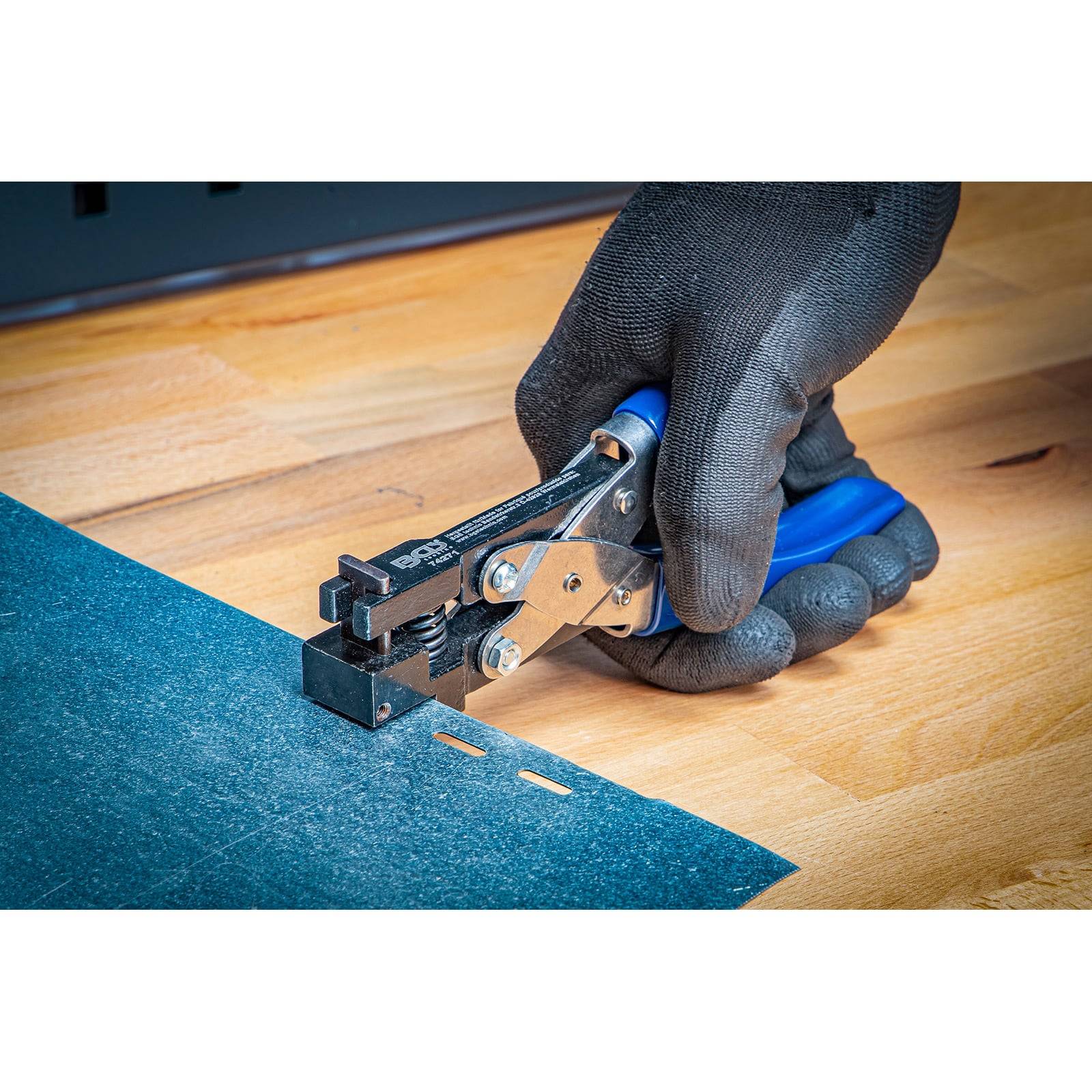 Eine mit einem Handschuh bekleidete Hand verwendet ein blau-silbernes Werkzeug, um Löcher in ein blaues Material auf einer Holzoberfläche zu stanzen.