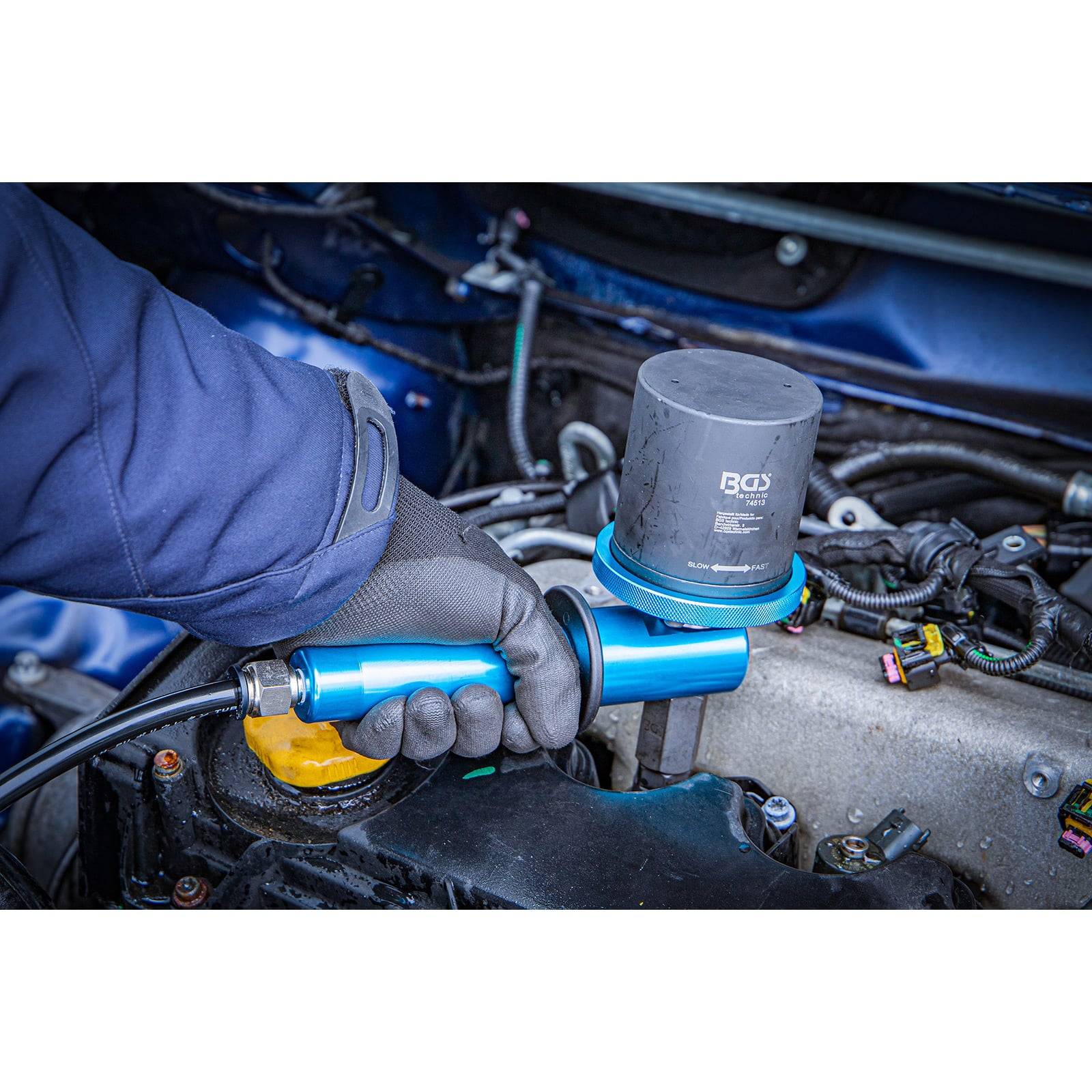 Eine Person benutzt ein Werkzeug, um ein Teil im Automotor zu installieren oder zu entfernen und trägt dabei Handschuhe als Sicherheitsmaßnahme. Die Motorkomponenten und Verkabelung sind sichtbar.