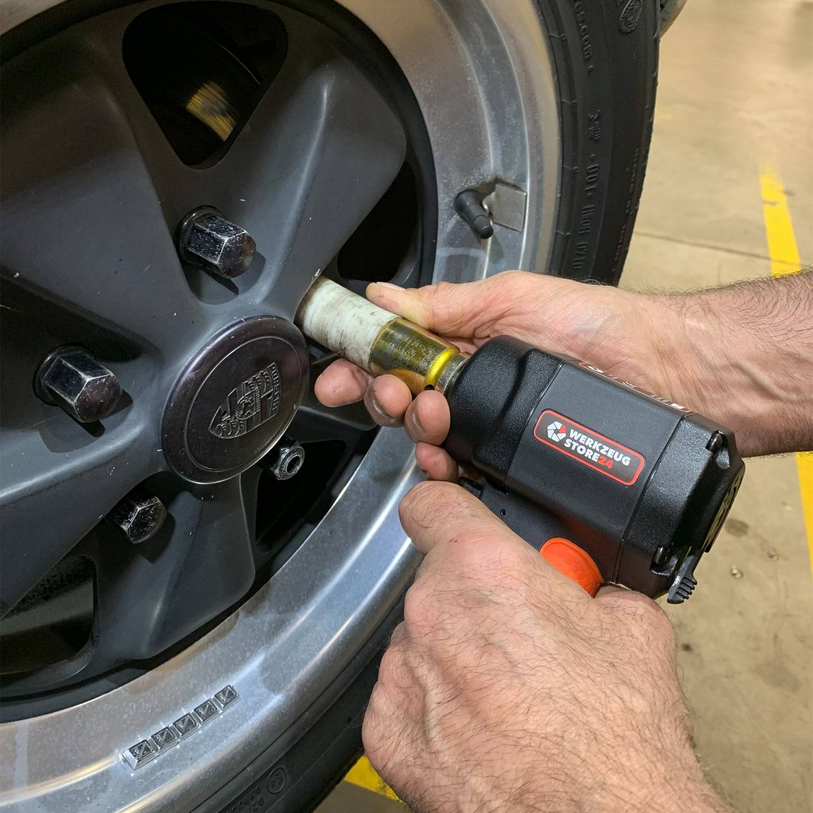 Ein Mechaniker verwendet ein Druckluftwerkzeug, um eine Radmutter an einem Autoreifens in einer Garagenumgebung anzuziehen. Das Rad hat fünf Radmuttern und ein sichtbares Autologo.
