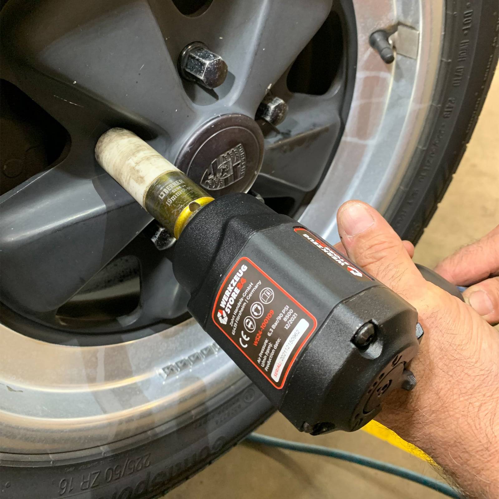 Schlagschrauber zieht eine Radmutter an einer Autofelge in einer Garagenumgebung, wobei eine menschliche Hand das Werkzeug hält.