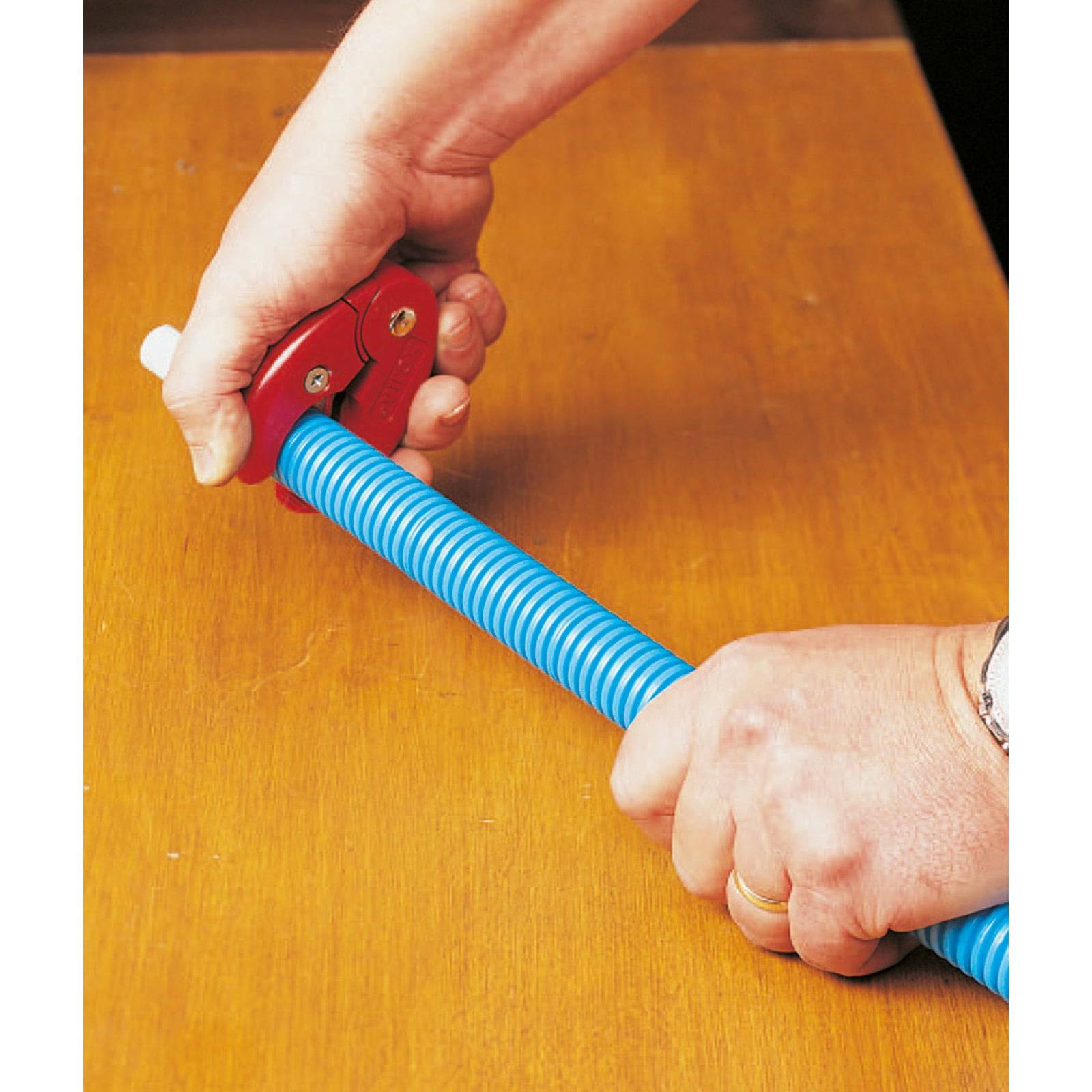 Zwei Hände benutzen einen roten Rohrabschneider, um ein blaues Wellrohr auf einer Holzoberfläche zu durchtrennen.