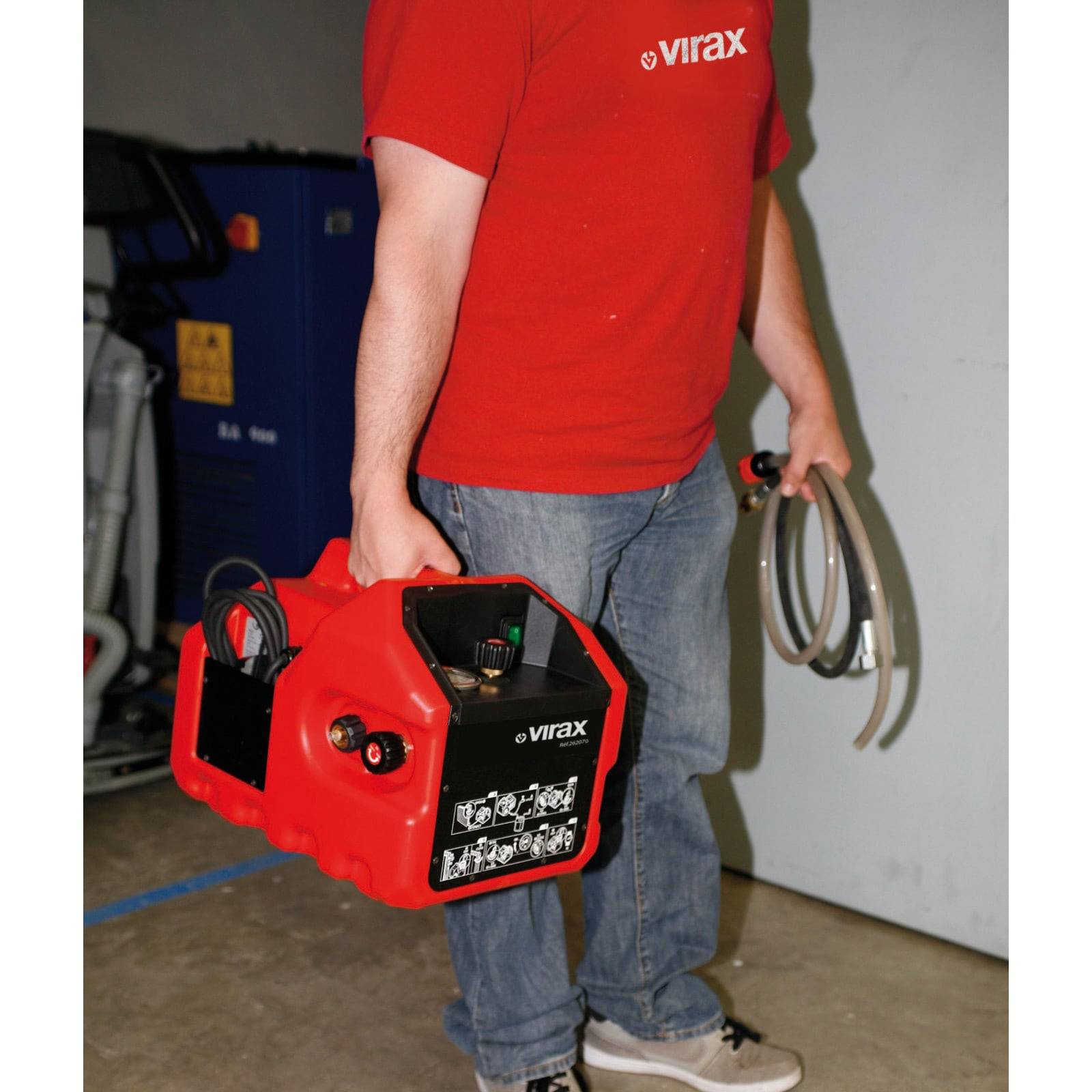Eine Person steht und hält eine rote Virax-Maschine und einen Schlauch, trägt ein passendes rotes Shirt mit 'Virax'-Branding, in einem industriellen Umfeld.