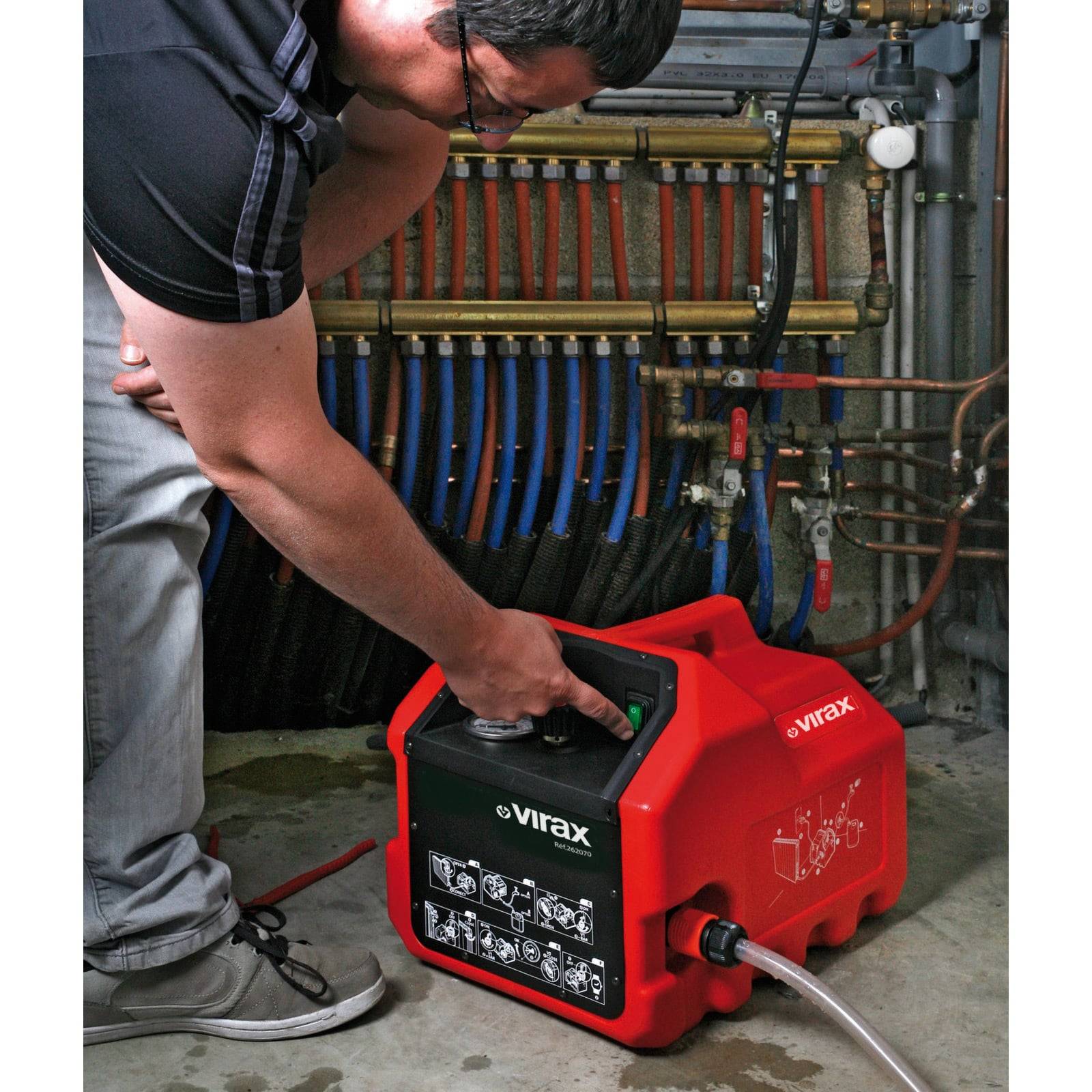 Eine Person bedient eine rote Virax-Druckprüfpumpe, die an blaue und rote Rohrleitungen an einer Wand angeschlossen ist. Das Gerät wird zum Überprüfen des Rohrdrucks verwendet.