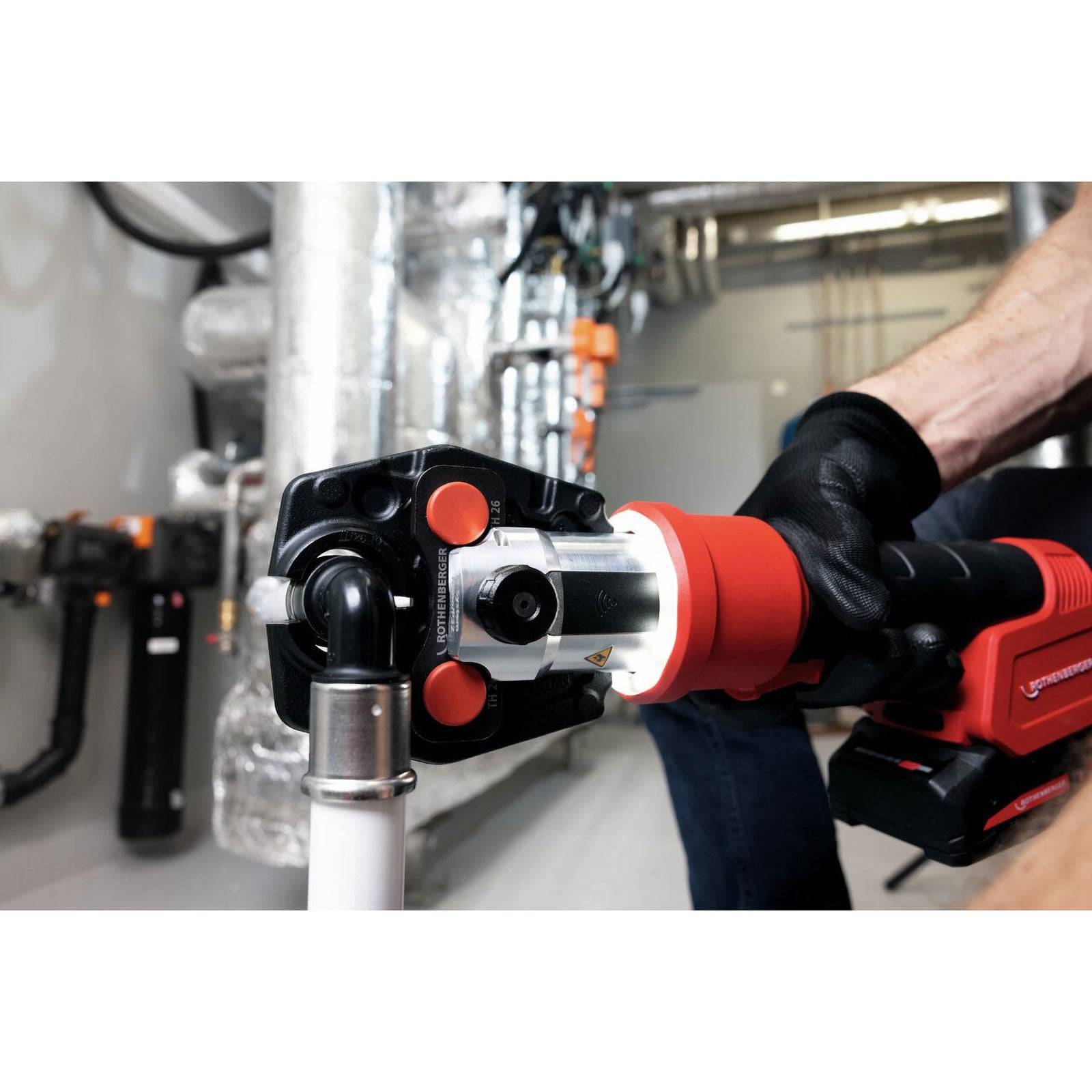 Eine Person benutzt ein rotes Werkzeug, um Metallrohrleitungen zu verbinden, wobei industrielle Ausrüstung im Hintergrund sichtbar ist.