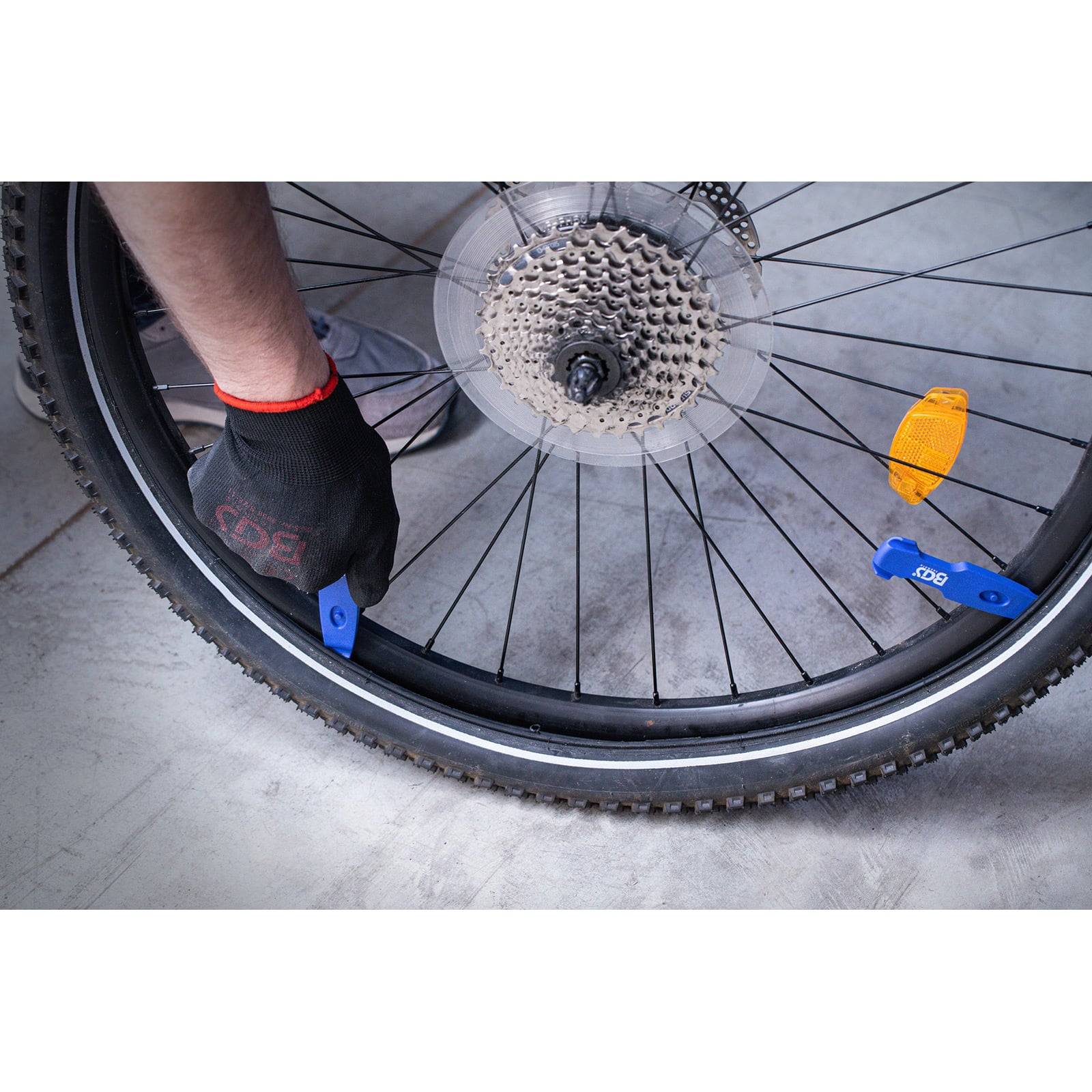 Eine Person benutzt ein Plastikwerkzeug, um einen Fahrradreifen von der Felge zu entfernen, wodurch die Zahnräder sichtbar werden. Die Person trägt einen schwarzen Handschuh mit roter Einfassung.