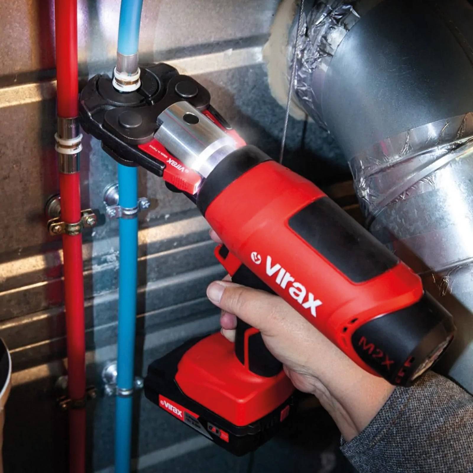 Eine Person benutzt ein rotes und schwarzes hydraulisches Werkzeug, um ein blaues Rohr neben einem roten zu klemmen. Das Werkzeug ist mit "virax"" beschriftet.