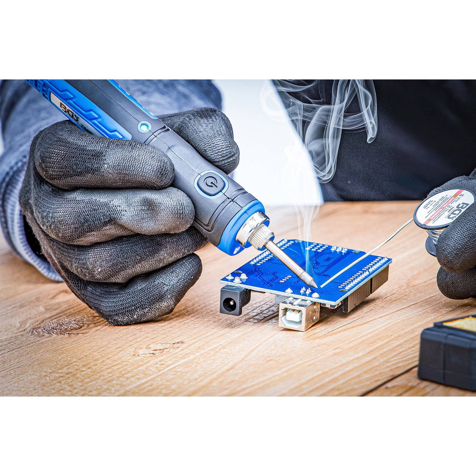 Eine Person lötet eine blaue Leiterplatte mit einem Lötkolben, wobei Rauch entsteht. Die Platine befindet sich auf einer Holzoberfläche.