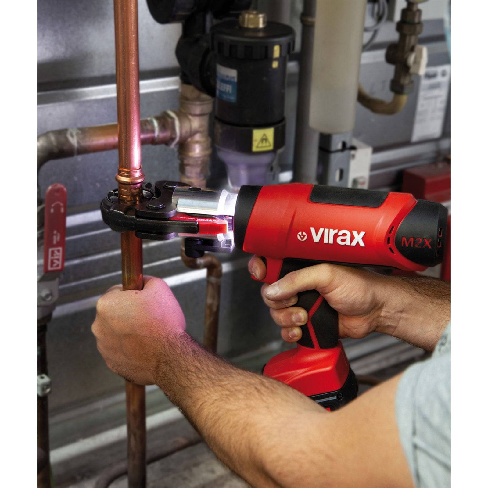 Person verwendet ein rotes Virax M2x Elektrowerkzeug zum Verpressvorgang von Verbindungen an Kupferrohren, wahrscheinlich während einer Sanitärinstallation oder Reparaturarbeit.