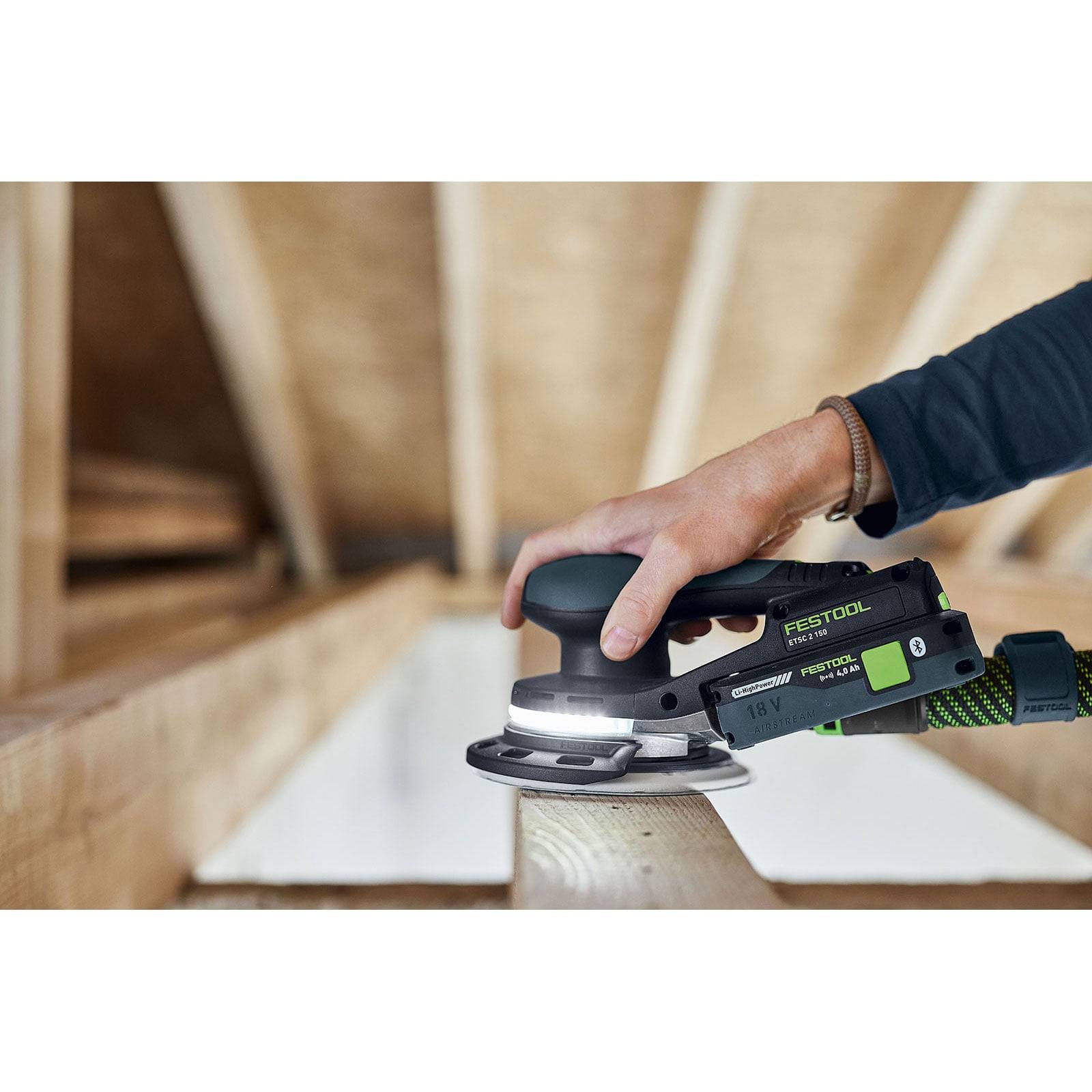 Eine Person schleift einen Holzbalken mit einem Festool-Schleifer in einem Dachbodenraum und veranschaulicht dabei die Werkzeuge, die in der Holz- oder Bauarbeit verwendet werden.