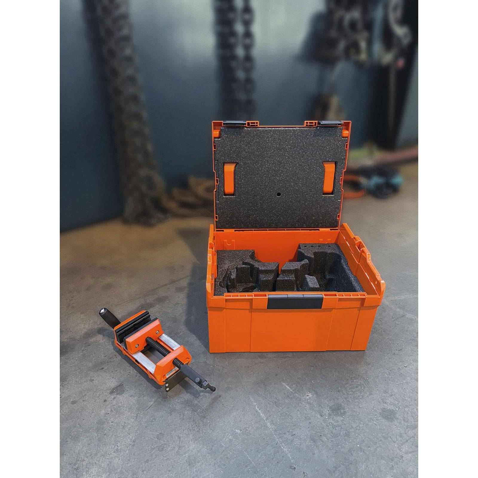 Eine orangefarbene robuste Aufbewahrungsbox ist geöffnet und zeigt Schaumstoffeinsätze im Inneren. Ein separates Werkzeug mit einem Griff liegt daneben auf dem Boden.
