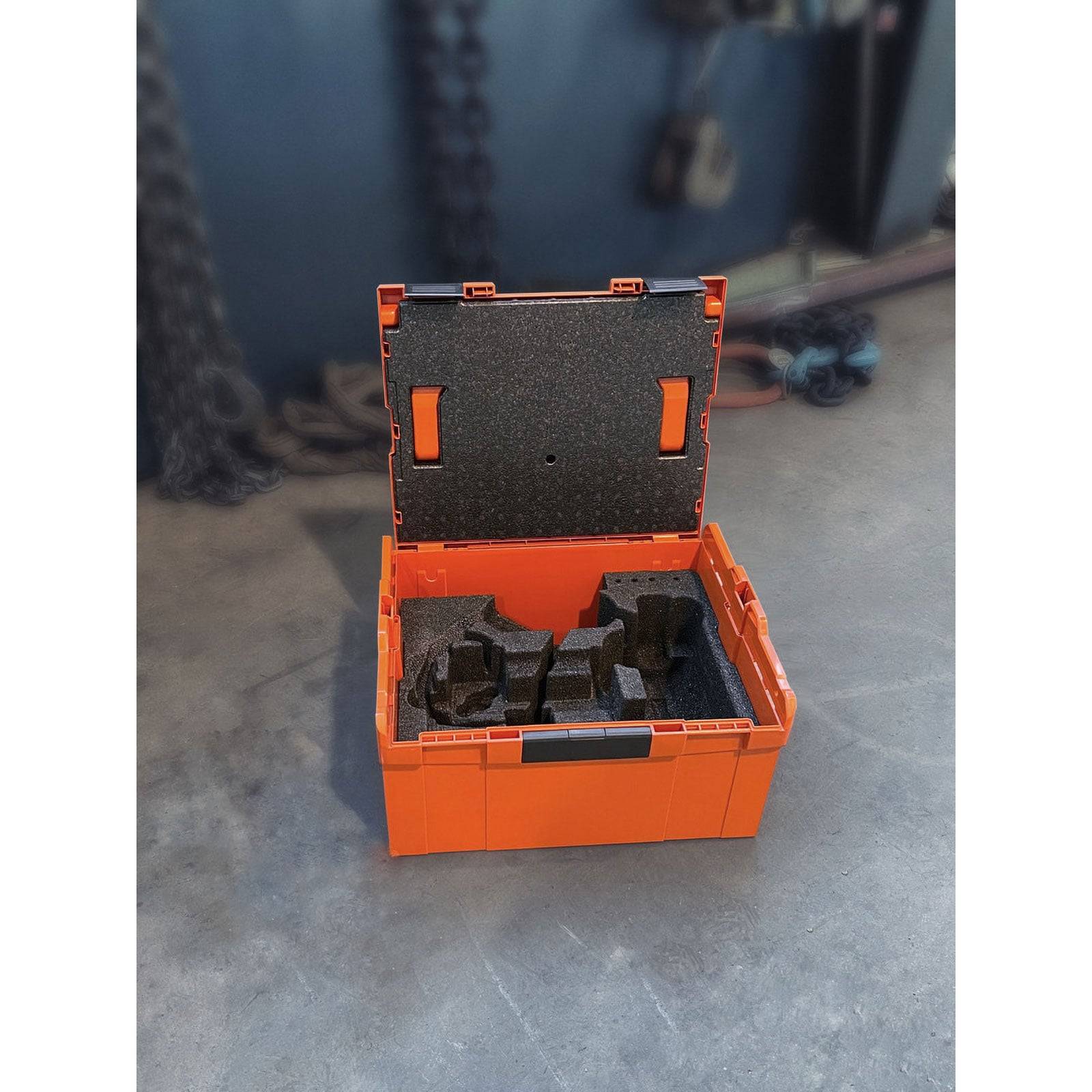 Eine offene orangefarbene Aufbewahrungsbox mit Schaumeinlagen steht auf dem Werkstattboden und ist von aufgerollten Ketten im Hintergrund umgeben.