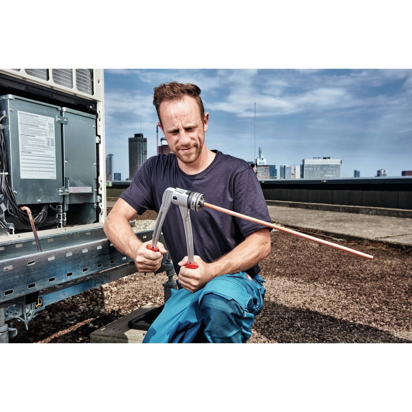 Eine Person kniet auf einem Dach und biegt ein Kupferrohr mit einem Werkzeug neben einer großen Klimaanlage, wobei eine Stadtlandschaft im Hintergrund zu sehen ist.
