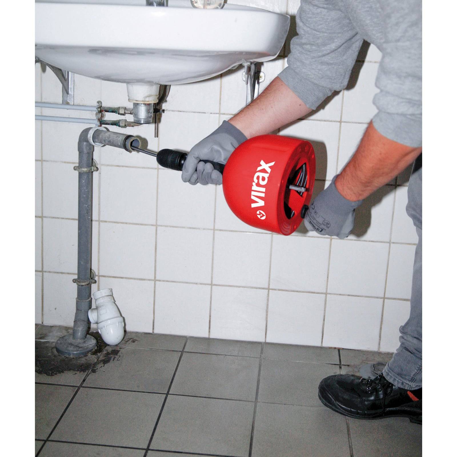 Eine Person benutzt einen roten manuellen Rohrreiniger der Marke 'Virax', um einen Abfluss unter einem Badezimmerwaschtisch zu entfernen. Der Boden ist gefliest, und die Klempnerrohre sind sichtbar.