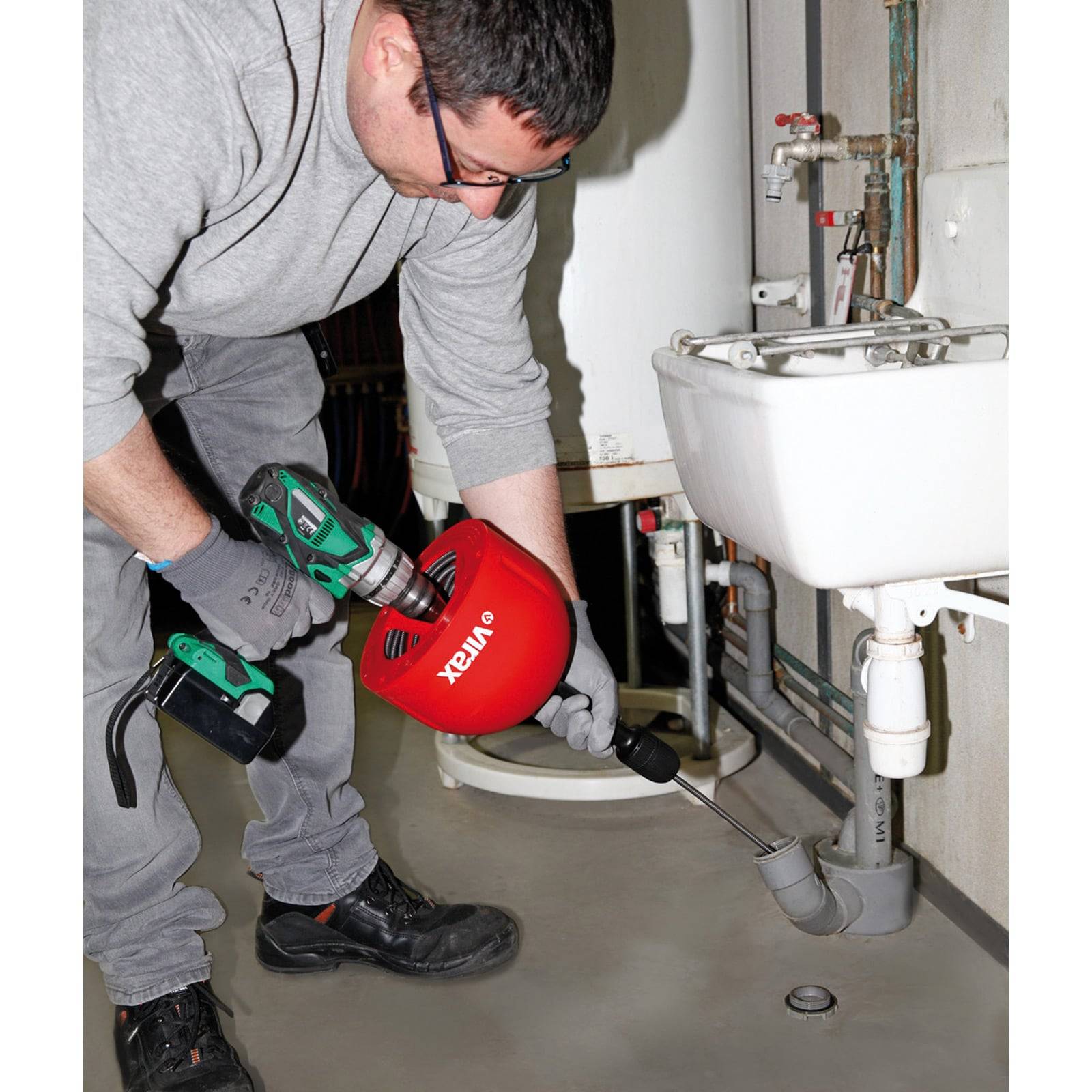 Eine Person benutzt ein Klempnerwerkzeug, um einen Abfluss in einem Badezimmer freizumachen. Die Szene umfasst einen Waschbecken, Rohre und einen Bohrmaschinenaufsatz.