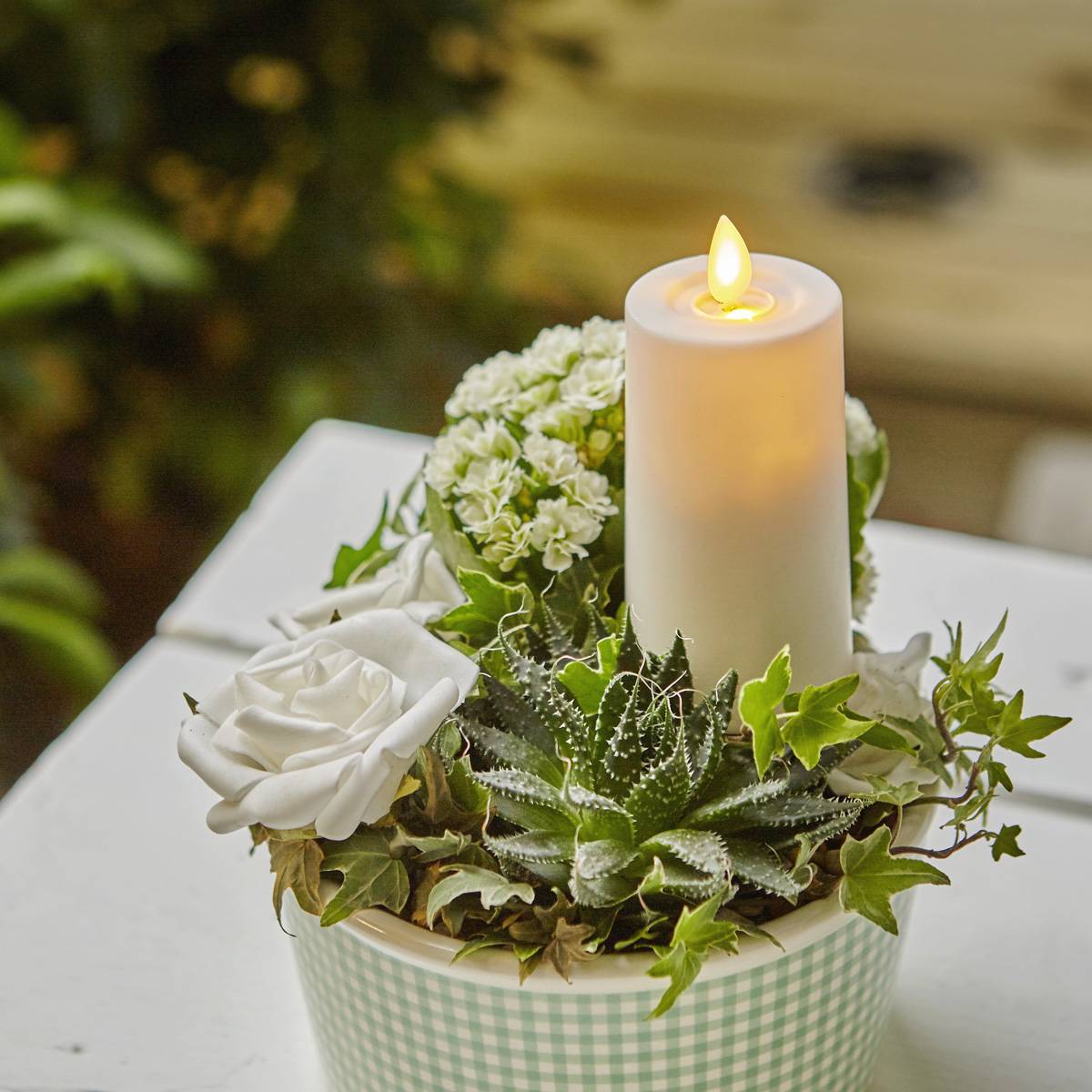 Eine weiße Kerze und verschiedene Blumen, darunter eine weiße Rose, in einem karierten Topf, der auf einer weißen Oberfläche steht, mit verschwommener Grünpflanzerie im Hintergrund.