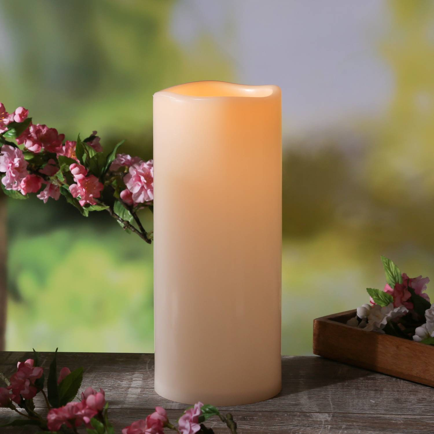 Eine brennende, hohe weiße Kerze auf einem Holztisch, umgeben von rosa Blumen, die eine ruhige und friedliche Atmosphäre schaffen.