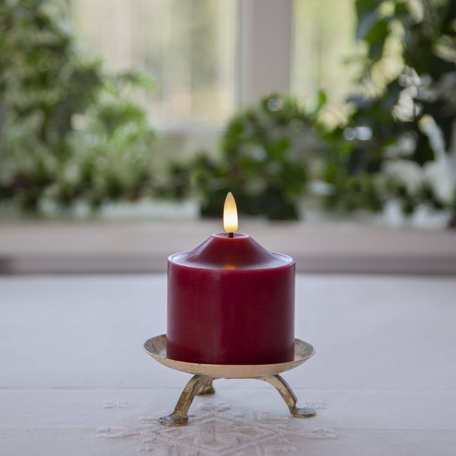 Rote Kerze, angezündet auf einem weißen Glasteller, zentriert auf einem Tisch mit verschwommenen grünen Pflanzen im Hintergrund.
