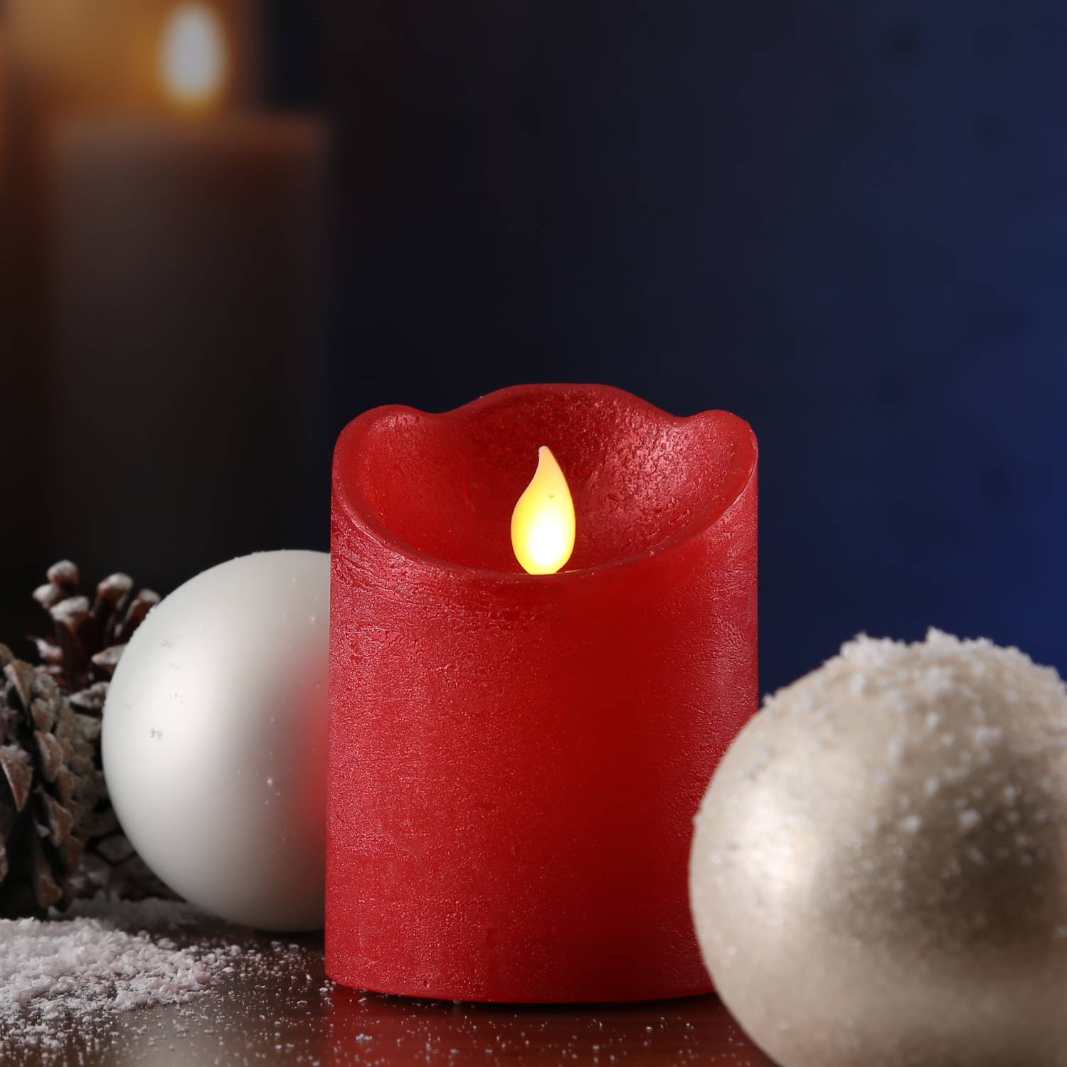 Eine rote Kerze leuchtet mit warmem Licht neben schneebedeckten Tannenzapfen und zwei weißen Ornamenten und schafft eine gemütliche Winteratmosphäre.