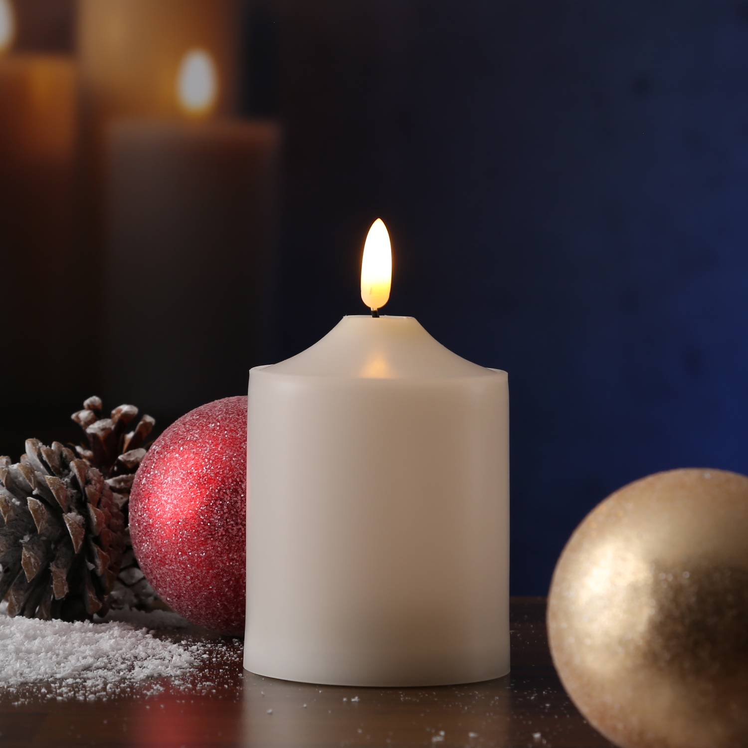 Eine brennende weiße Kerze, umgeben von roten und goldenen Ornamenten und einer Tannenzapfen auf einer Holzoberfläche, vor einem dunklen, unscharfen Hintergrund.