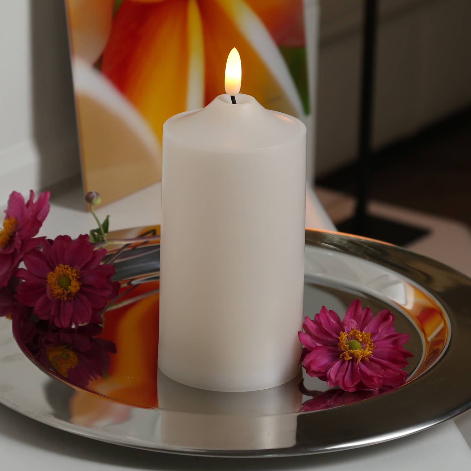 Eine leuchtende weiße Kerze auf einem reflektierenden silbernen Tablett, umgeben von rosa Blumen, die eine warme und ruhige Atmosphäre schaffen.