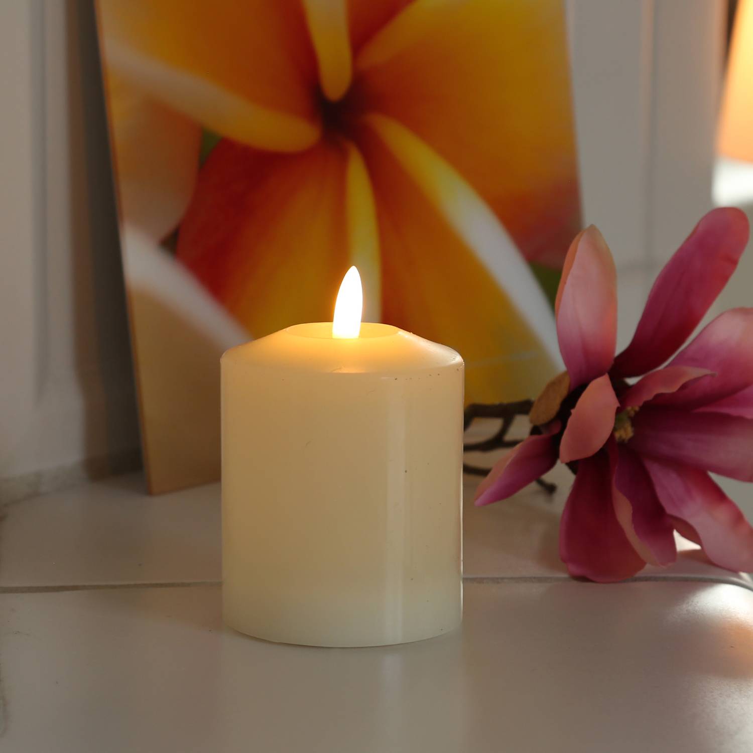 Eine brennende weiße Kerze neben einer rosa Blume und einem lebhaften, abstrakten Blumengemälde im Hintergrund.