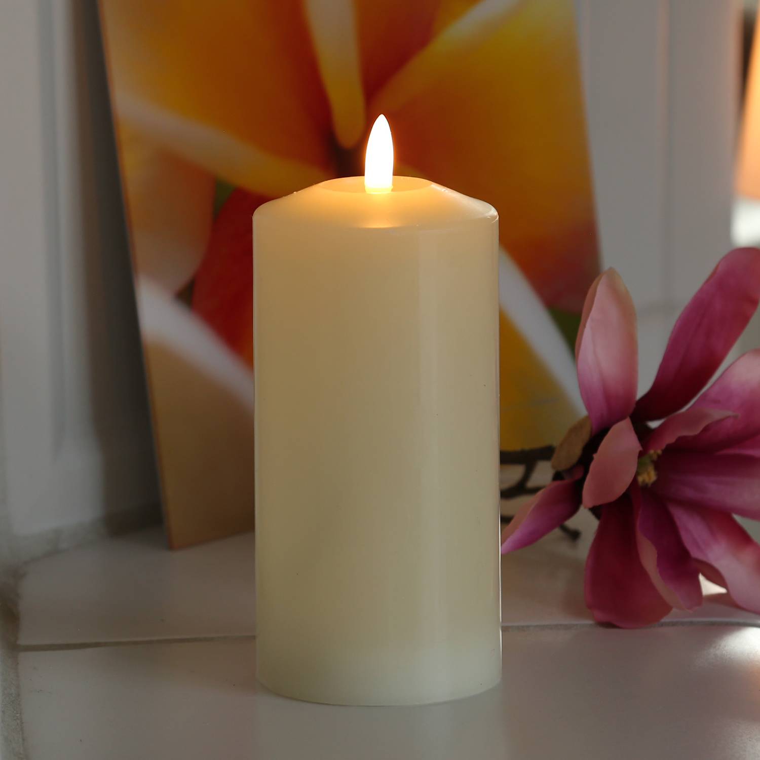 Eine brennende elfenbeinfarben Kerze auf einer weißen Oberfläche, daneben eine rosa Blume, vor einem verschwommenen, bunten Blumenhintergrund.