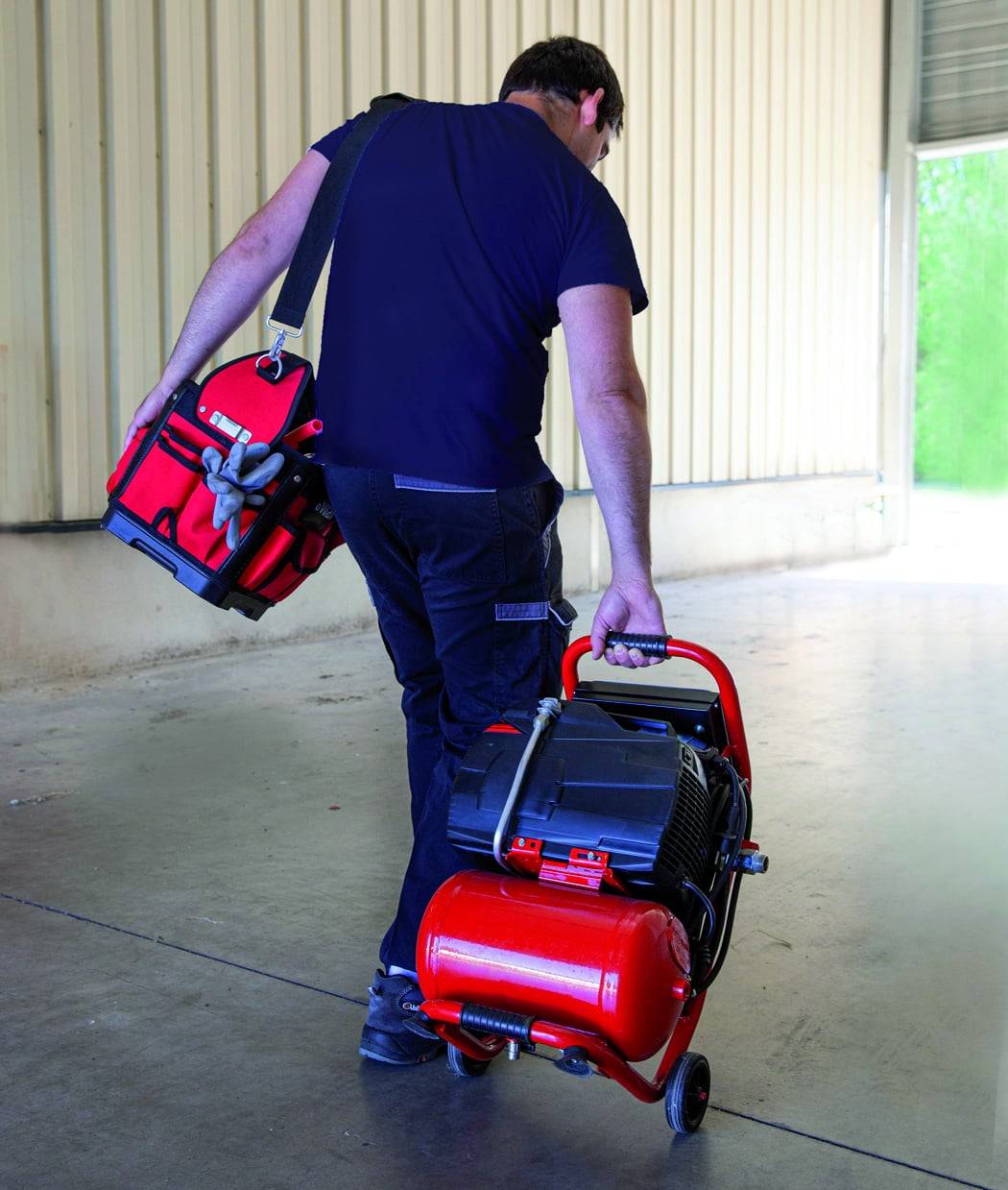 Eine Person, gekleidet in Schwarz, trägt eine rote Werkzeugkiste auf der Schulter und einen roten Kompressor in der anderen Hand, und geht an einem Metallgebäude vorbei.