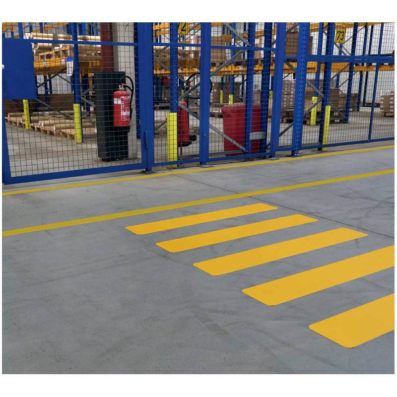 Ein Lagerbereich mit einem gelben Fußgängerüberweg auf einem Betonboden, Metallzaun, Feuerlöscher und Lagerregalen im Hintergrund.