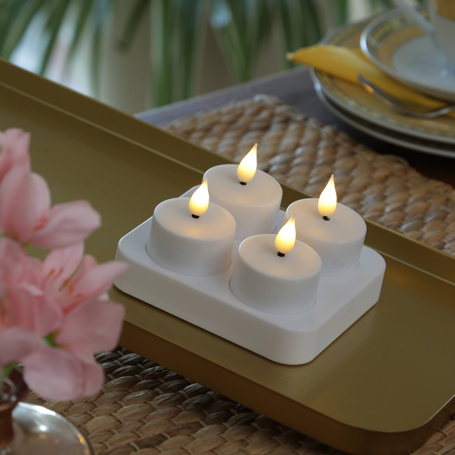 Vier leuchtende weiße flammenlose Kerzen auf einem weißen Tablett auf einem goldenen Tisch, mit rosa Blumen im Vordergrund und Besteck im Hintergrund.