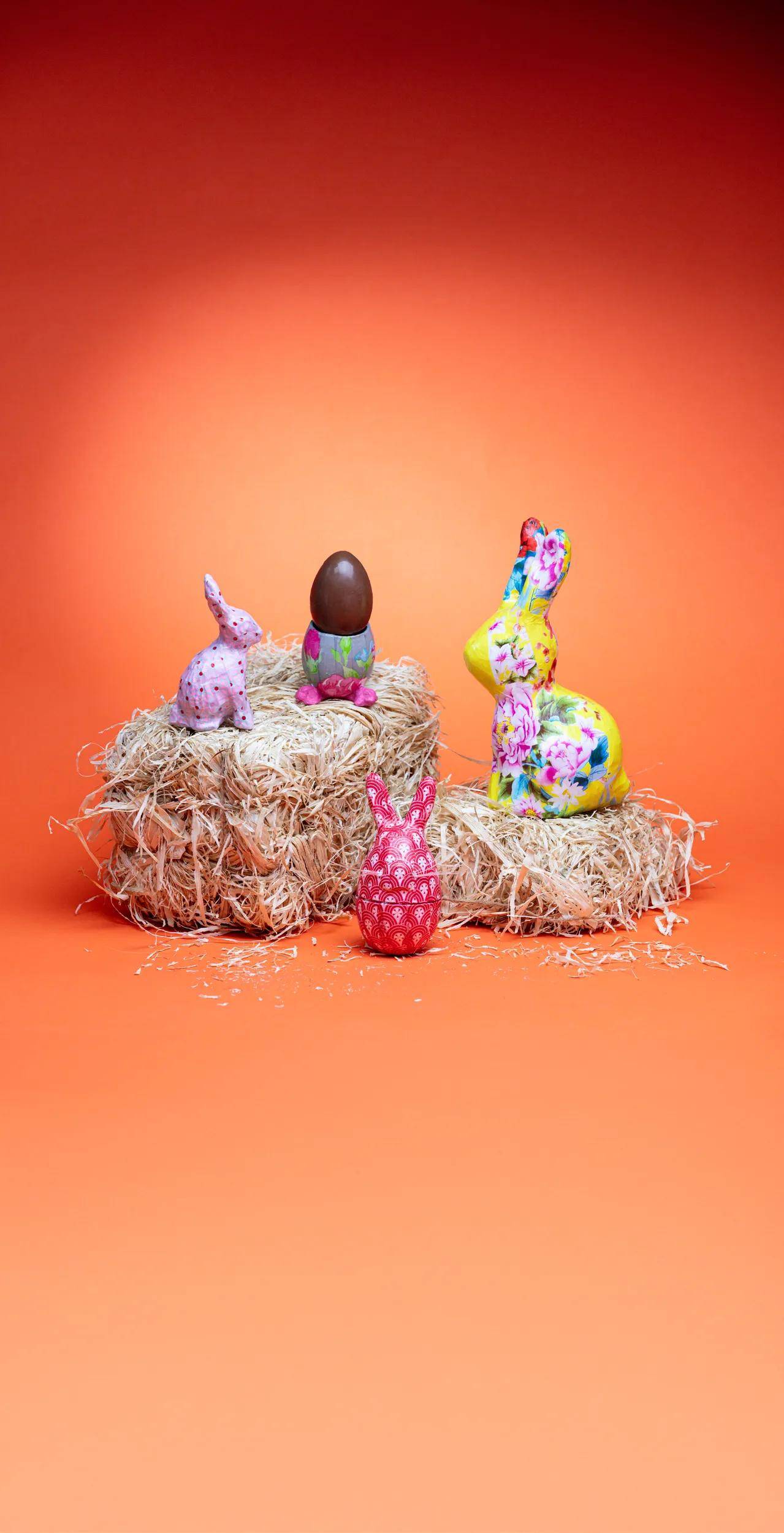 Drei dekorative Kaninchenfiguren, eine gemustert und eine mit einem Ei als Kopf, sitzen auf Heu vor einem orangefarbenen Hintergrund.