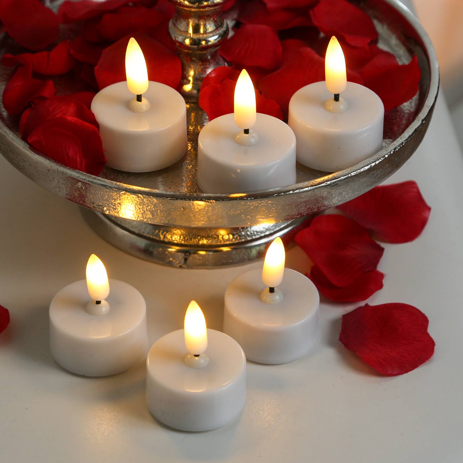Fünf brennende Teelichter, die um einen Metallständer mit Rosenblütenblättern angeordnet sind und eine warme, romantische Atmosphäre schaffen.