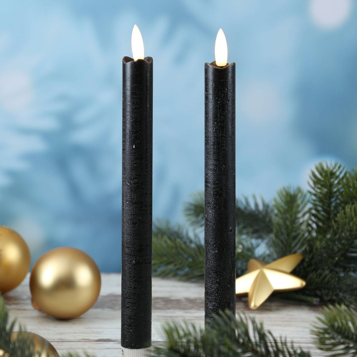 Zwei brennende schwarze Kerzen, umgeben von Tannenzweigen und goldenen Weihnachtsschmuck auf einem Holztisch, vor einem weichen blauen Hintergrund.