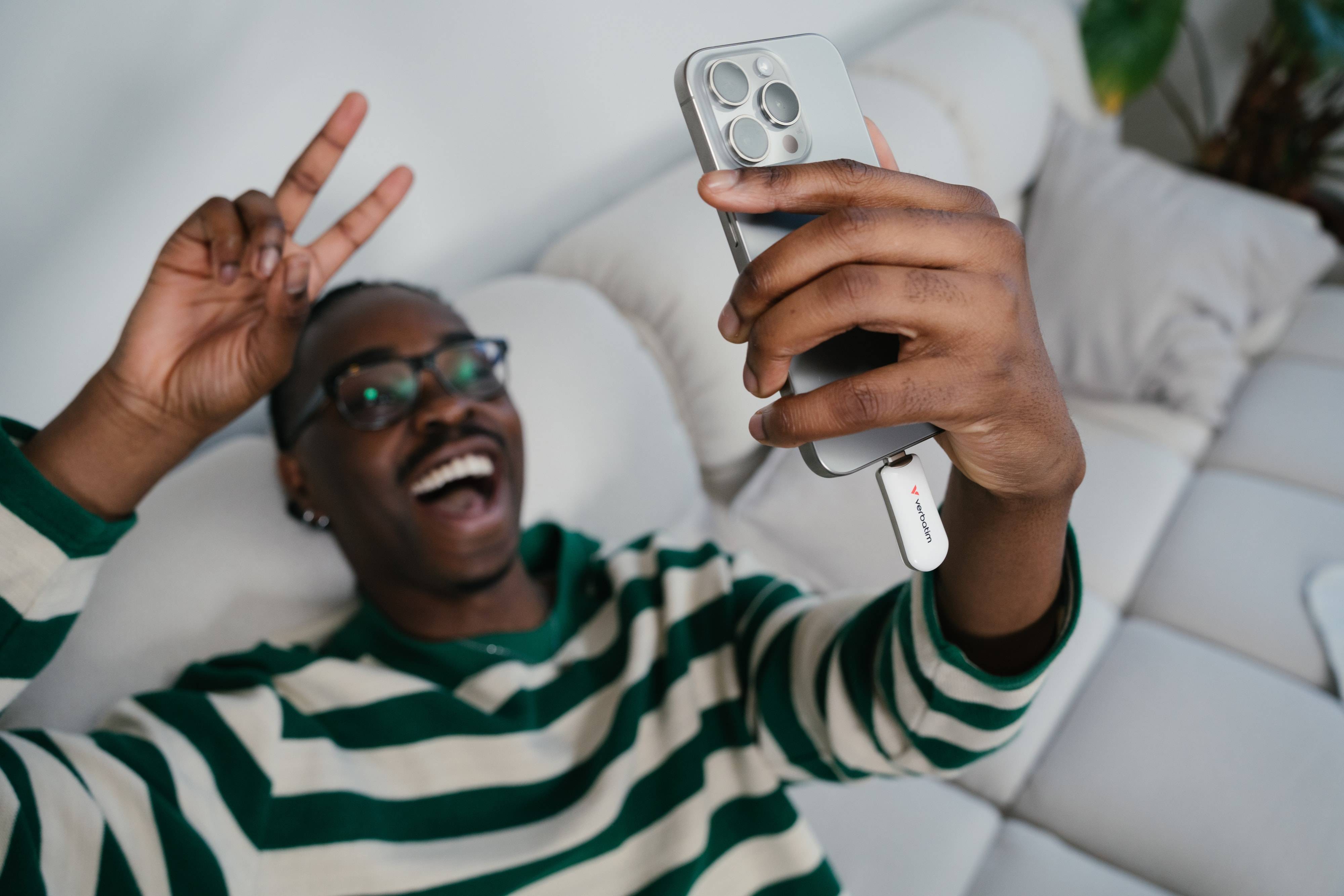Lächelnder Mann auf einem Sofa macht ein Selfie mit einem Smartphone und zeigt ein Friedenszeichen.