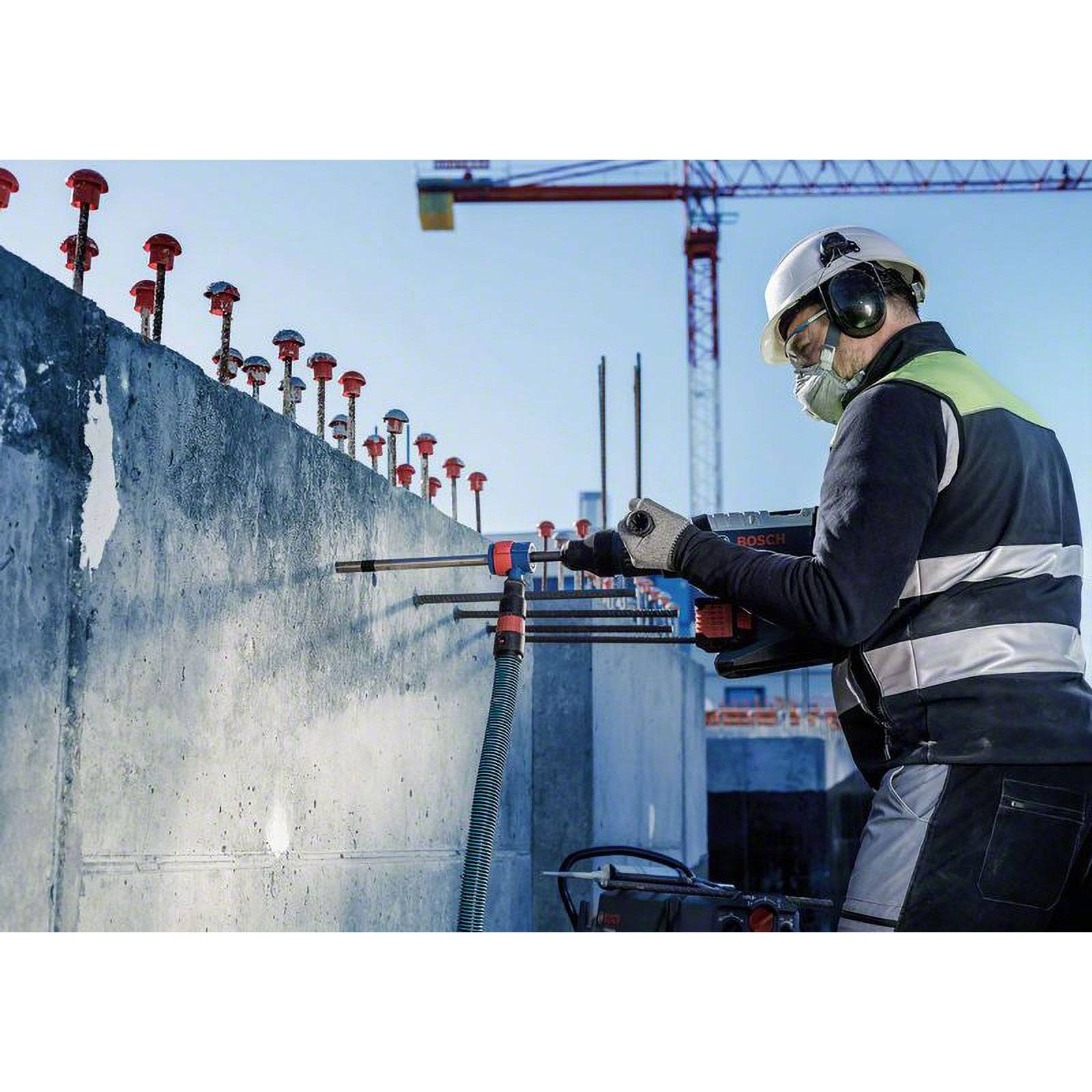 Ein Bauarbeiter in einer Sicherheitsweste und Helm benutzt ein Bohwerkzeug an einer Betonwand auf einer Baustelle mit Kränen im Hintergrund.