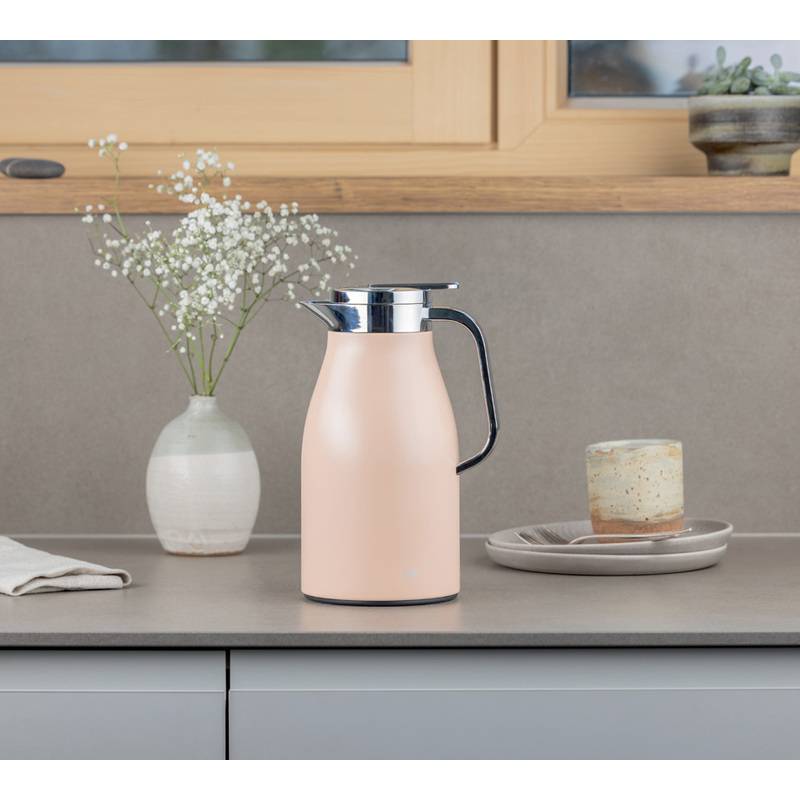 Eine rosa Thermoskanne mit Henkel steht auf einer Küchenarbeitsplatte neben einer Vase mit weißen Blumen und einer Tasse auf einem Unterteller.