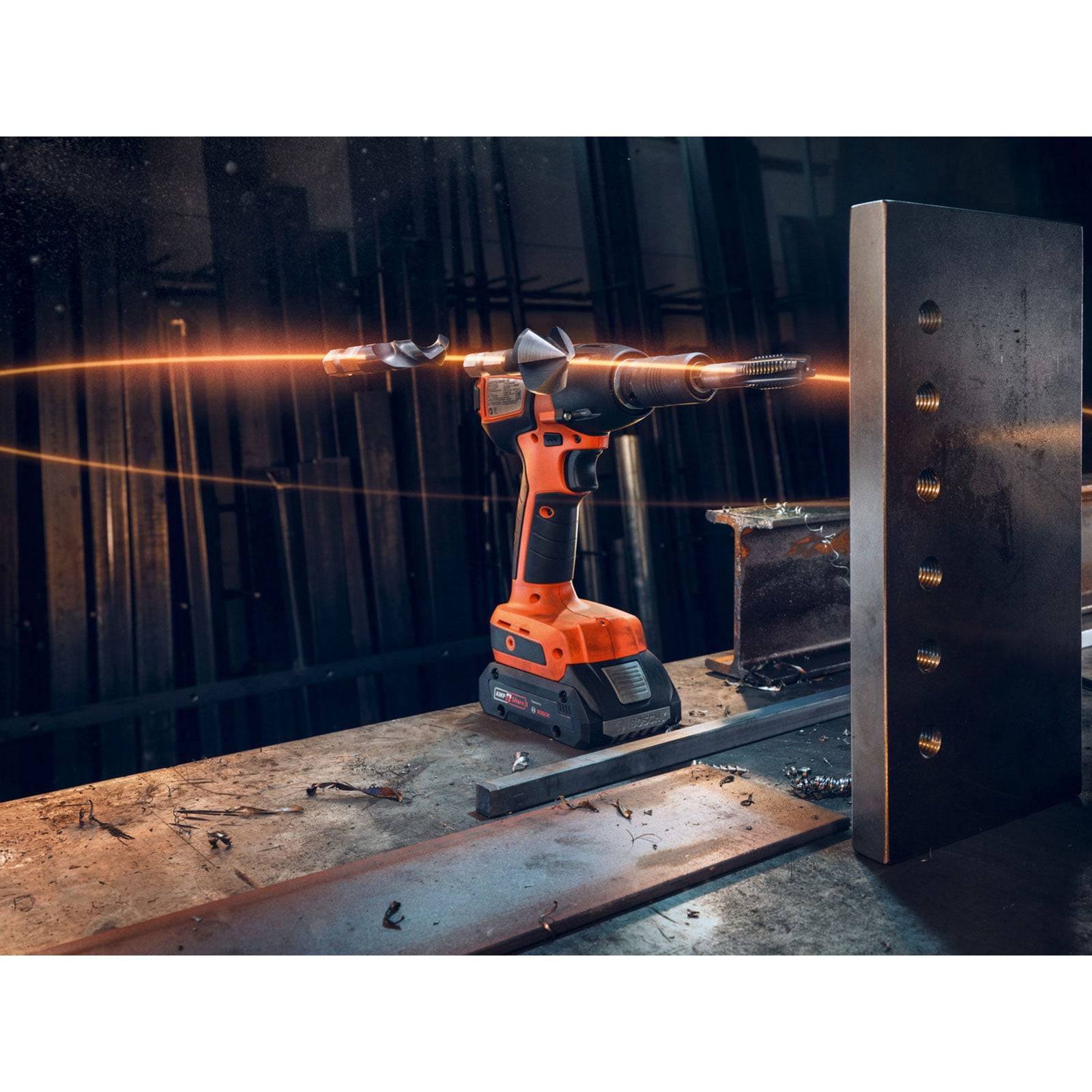 Eine Bohrmaschine mit orangefarbenem Gehäuse liegt auf einer Werkbank in einer dämmrig beleuchteten Werkstatt. Eine Metallplatte mit Löchern befindet sich in der Nähe.