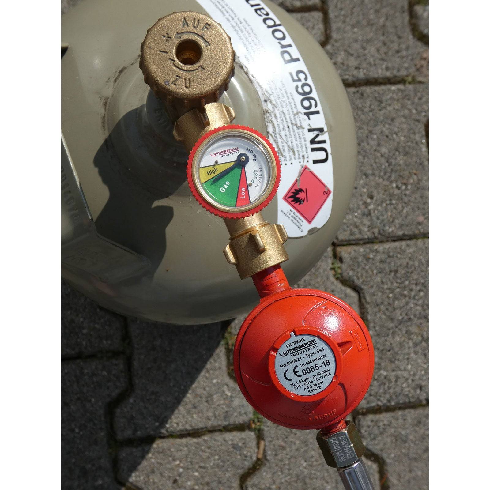 Propan-Gasflasche mit Druckmesser und rotem Regler, gekennzeichnet mit UN 1965 Propan. Warnhinweis weist auf entzündliche Inhalte hin.