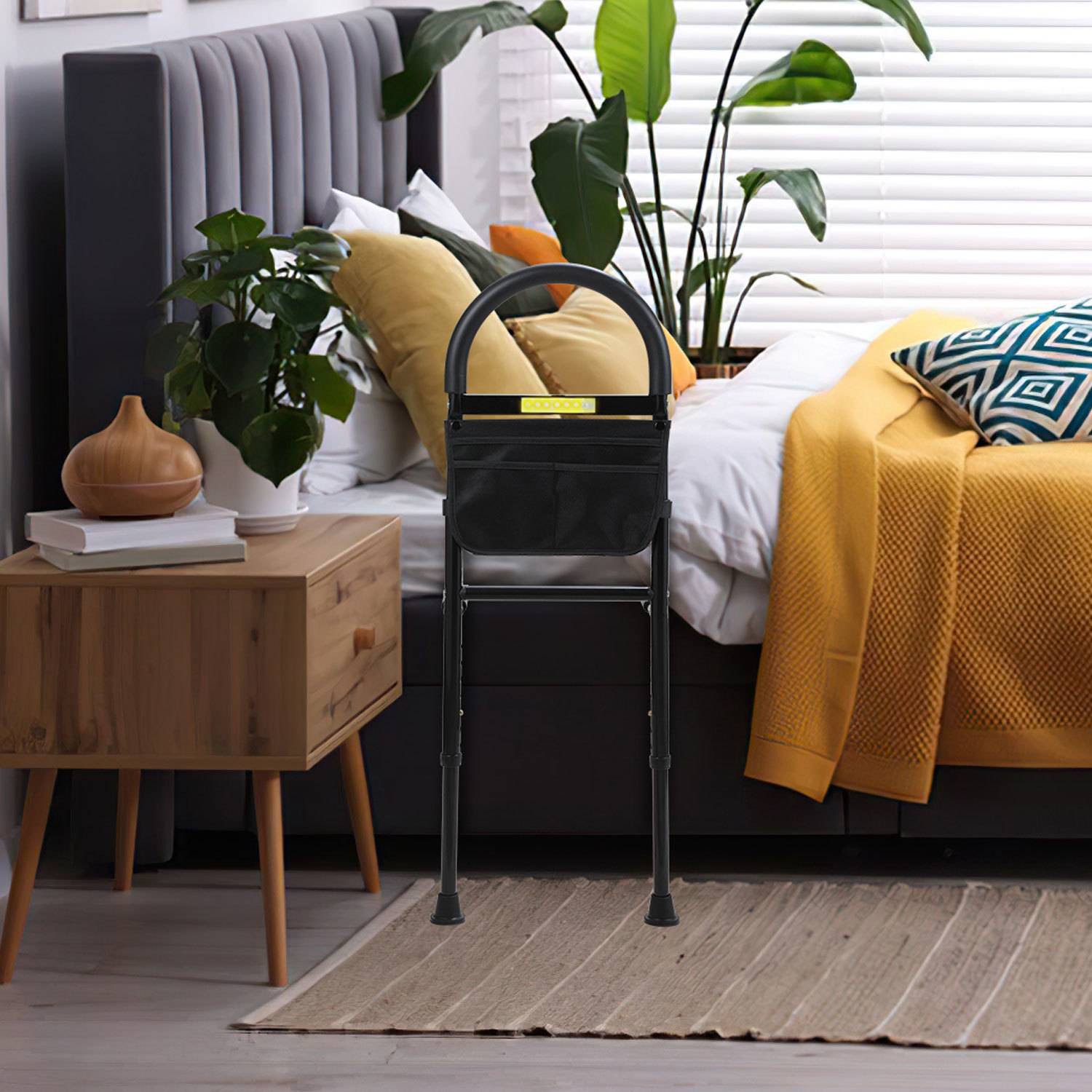 Ein schwarzer klappbarer Tritthocker steht in einem modernen Schlafzimmer neben einem Bett mit gelber Bettwäsche, einem Nachttisch und Topfpflanzen.
