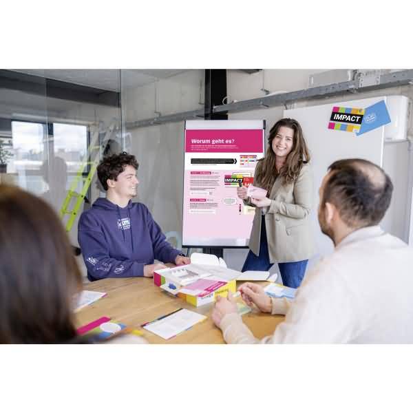 Frau präsentiert ein Plakat einer kleinen Gruppe in einem Konferenzraum, während Menschen um einen Tisch sitzen und zuhören sowie diskutieren.