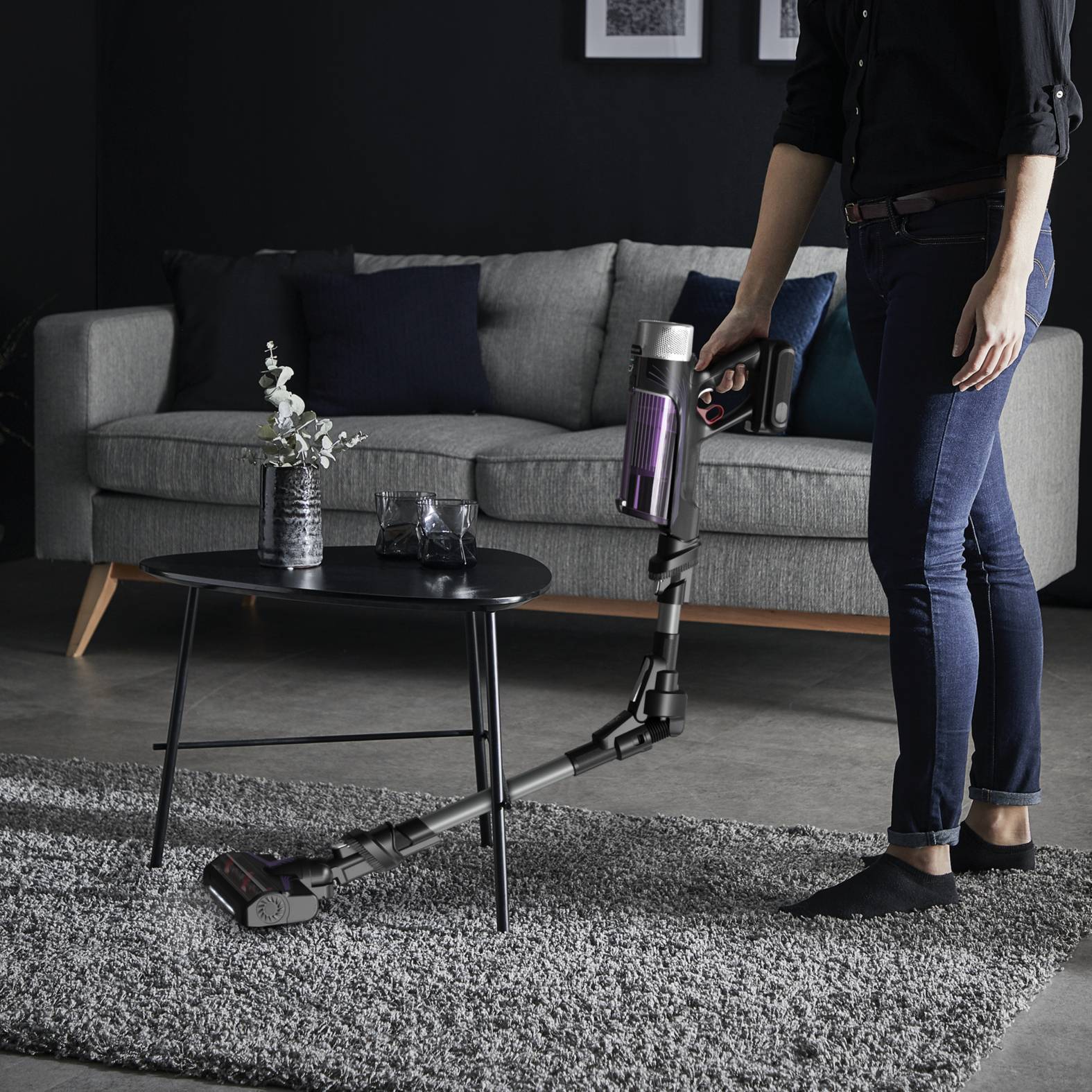 Eine Person, die mit einem kabellosen Stabstaubsauger ein Wohnzimmer neben einem grauen Sofa und einem Couchtisch absaugt.