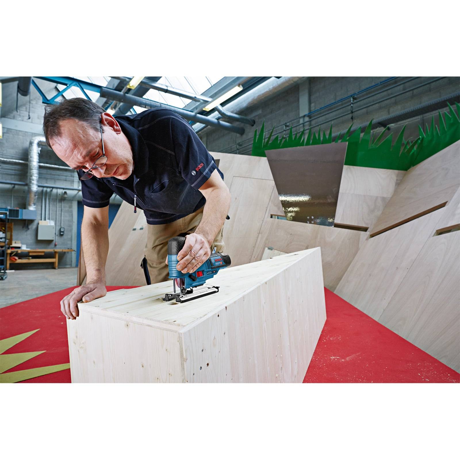 Eine Person verwendet ein Elektrowerkzeug, um ein Stück Holz in einer Werkstatt mit großen geometrischen Holzstrukturen im Hintergrund zu schneiden.