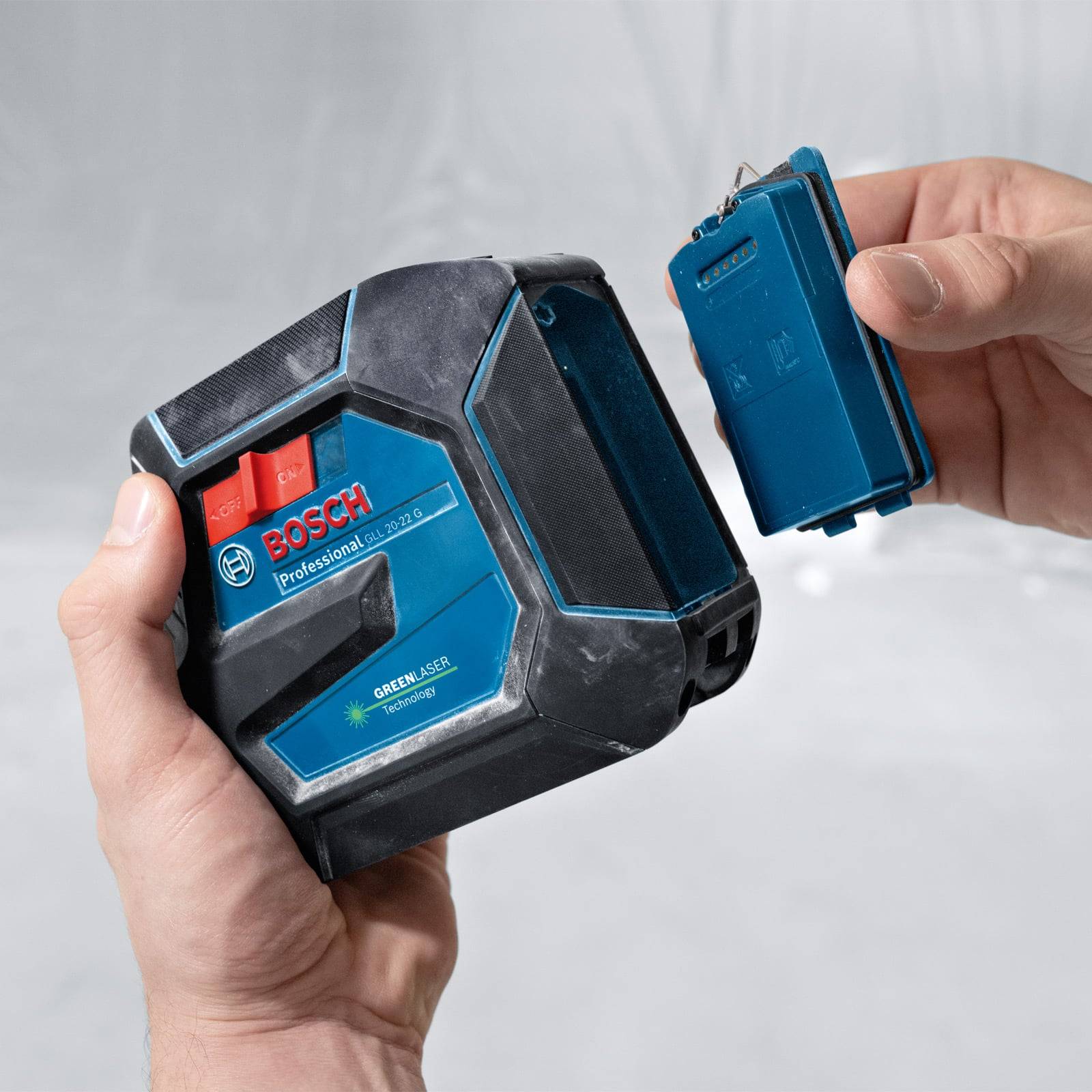 Zwei Hände, die ein professionelles Bosch-Lasernivelliergerät halten, wobei der Akku abgenommen wird, was das Produktdesign und die Verwendung hervorhebt.