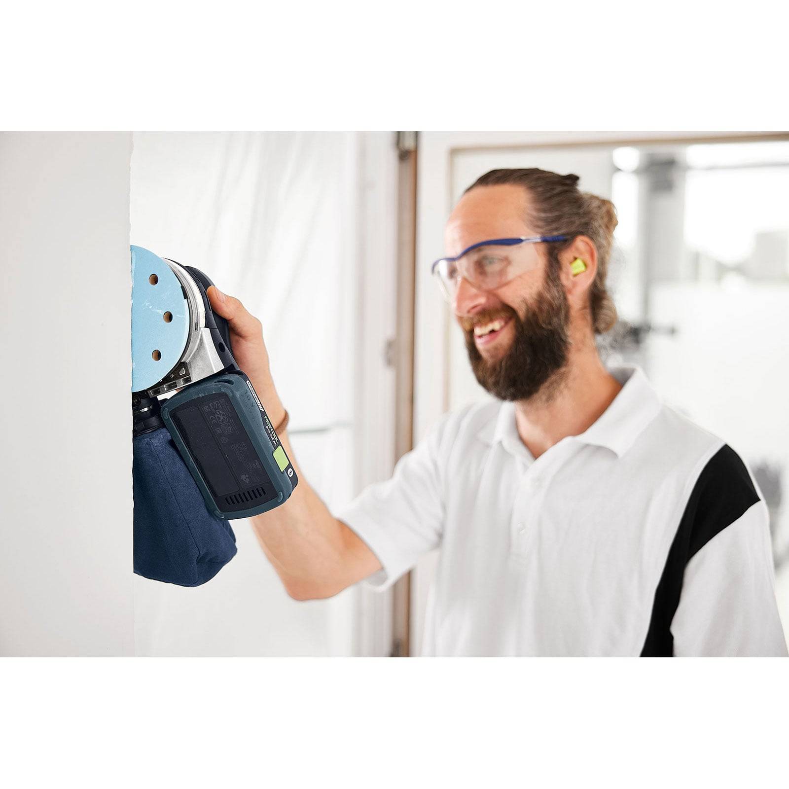 Ein Mann mit Sicherheitsbrille und Gehörschutz nutzt einen elektrischen Schleifer an einer weißen Wand und lächelt dabei bei seiner Arbeit.