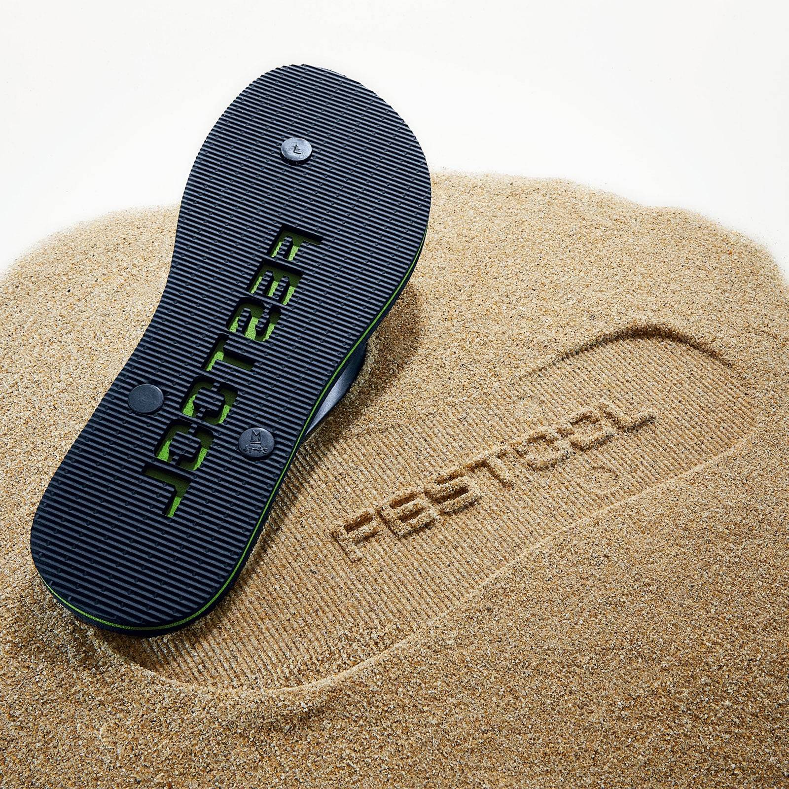 Ein Paar Sandalen mit der Aufschrift 'FESTOOL' auf den Sohlen hinterlässt einen Abdruck im Sand. Der Hintergrund ist sandig und hebt das Branding des Schuhs hervor.
