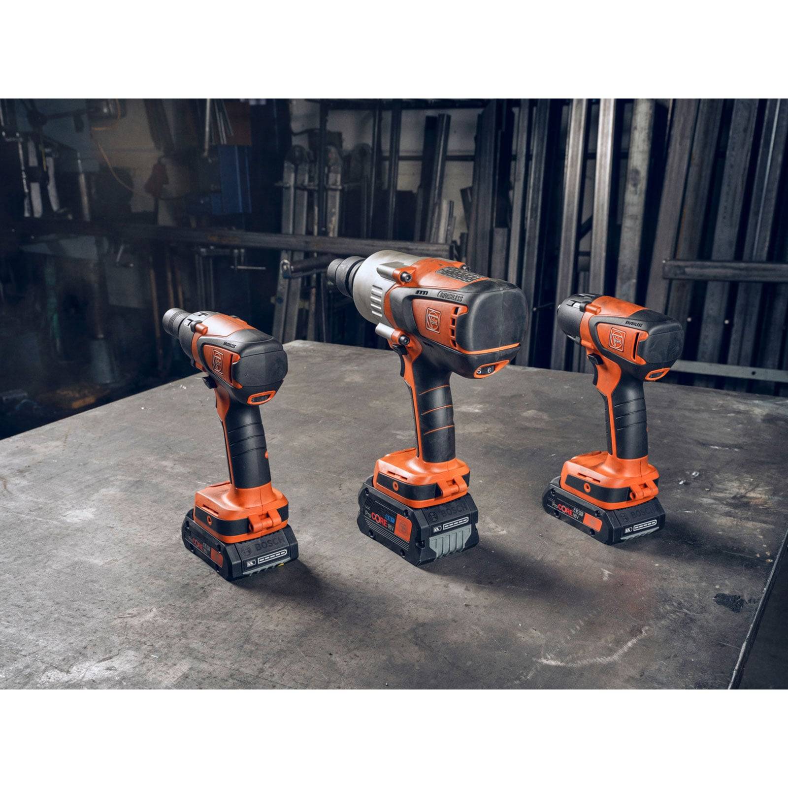 Drei orange und schwarze kabellose Akkuschrauber sind auf einem Werkstatttisch platziert, mit industriellen Regalen und Werkzeugen im Hintergrund.